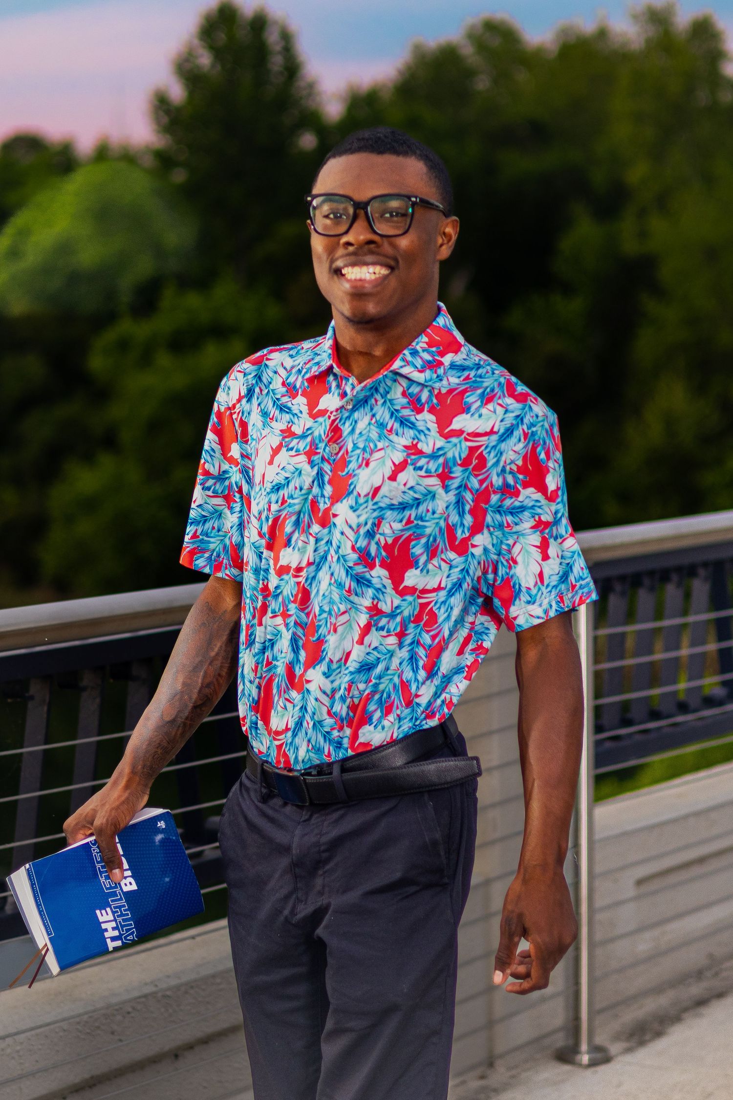 Smiling young man in colorful floral shirt and glasses holding a book outdoors on a bridge at dusk.