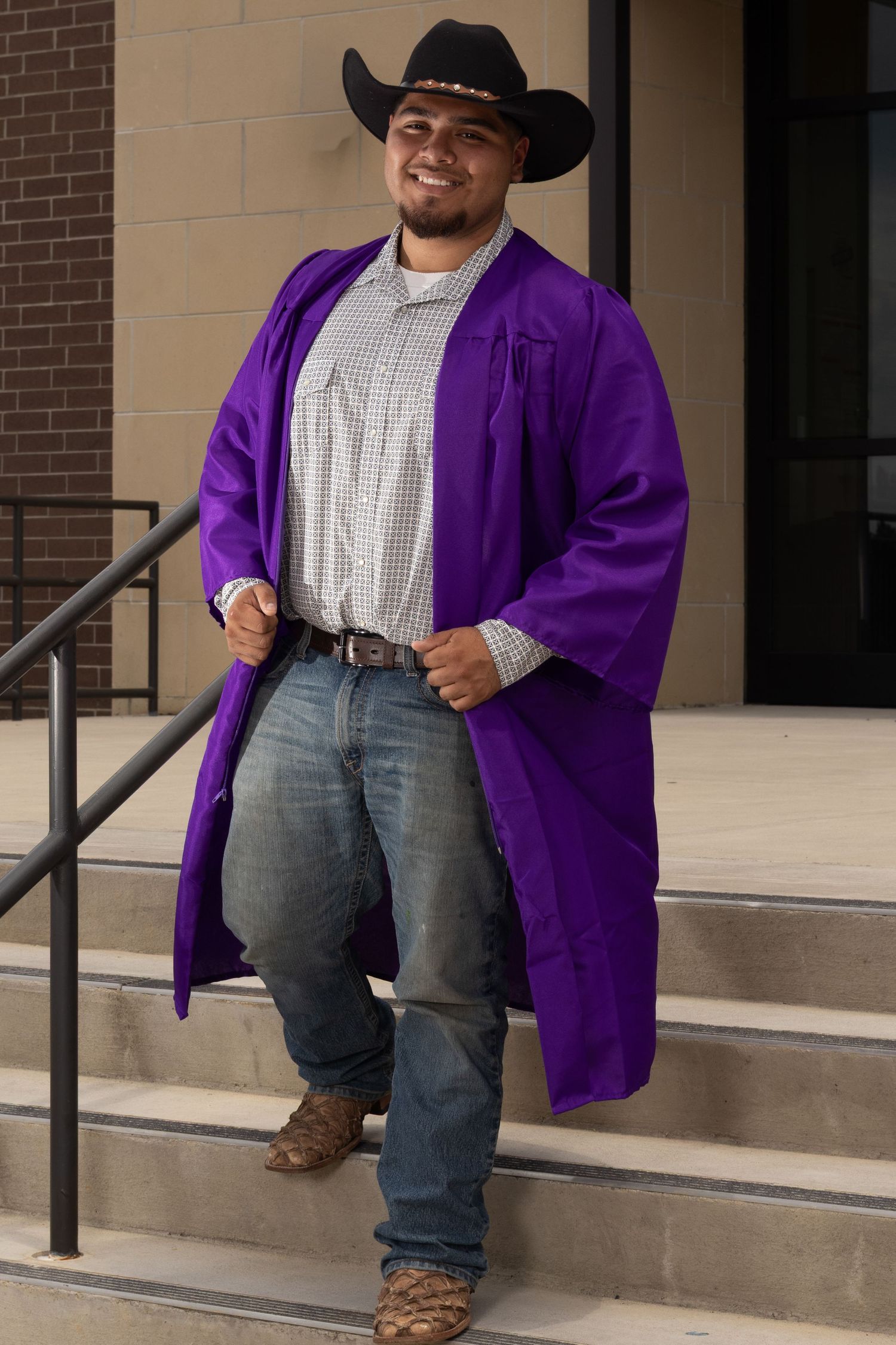 Smiling graduate in purple graduation robe and black cowboy hat poses on steps in jeans and boots.
