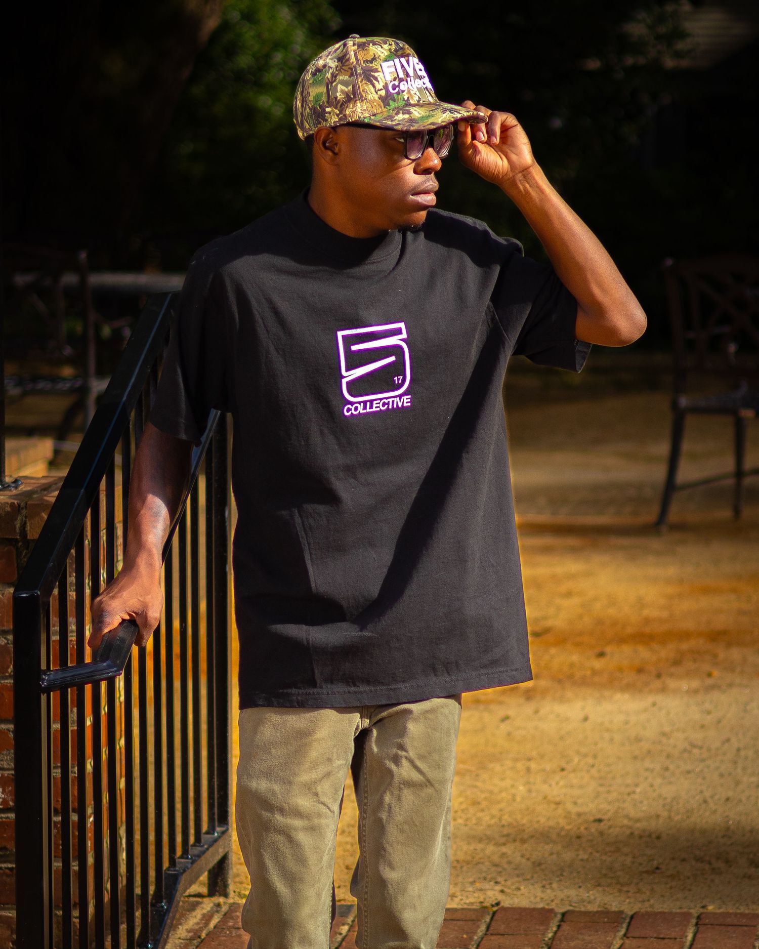 Young man wearing black graphic tee and camo cap poses confidently outdoors near a railing at night.