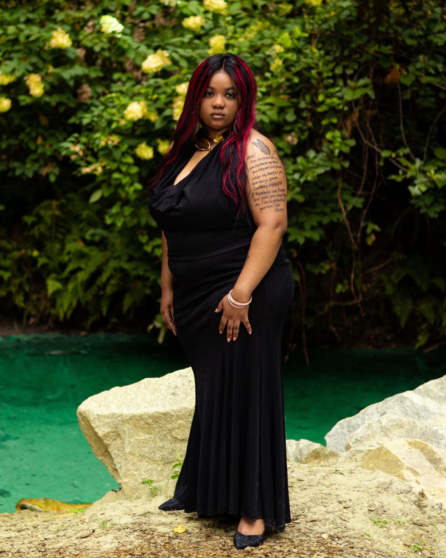 Woman in black maxi dress with red hair and arm tattoo poses on rocks near green water with lush foliage.