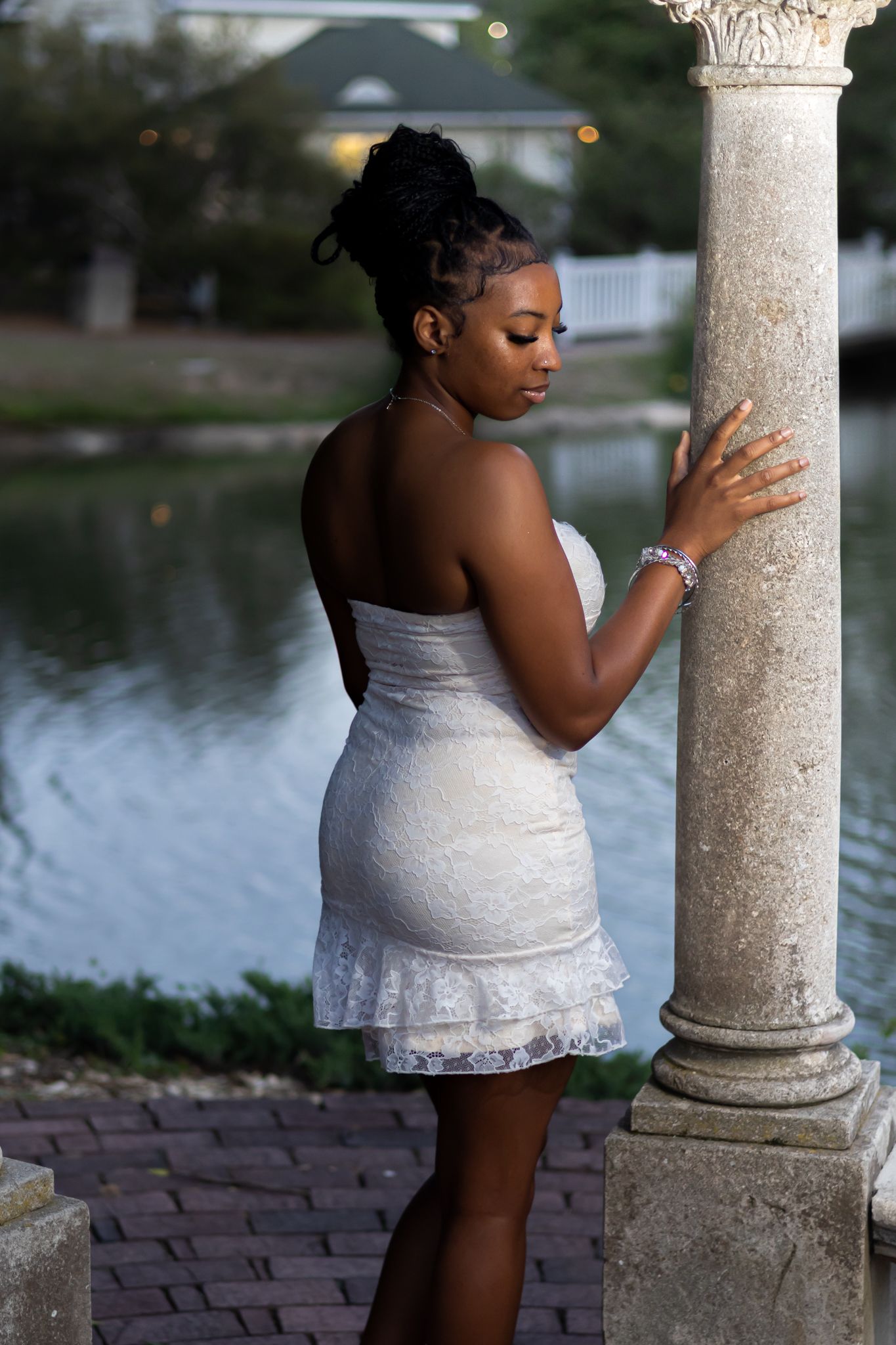 Young Black woman in white lace mini dress poses gracefully by stone column near waterfront at dusk.