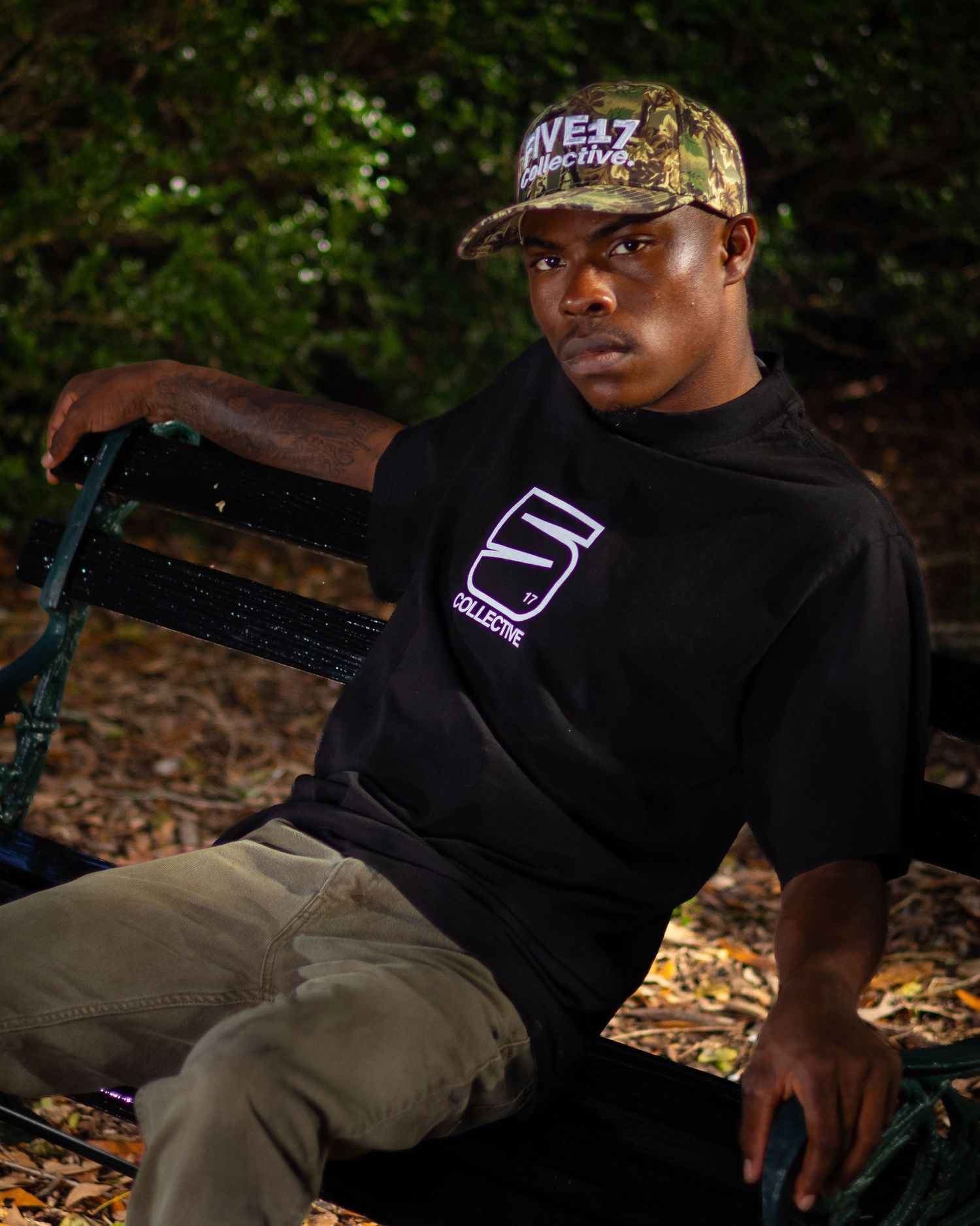 Young man in black graphic tee and camo cap sits on a bench outdoors at night, posing confidently.