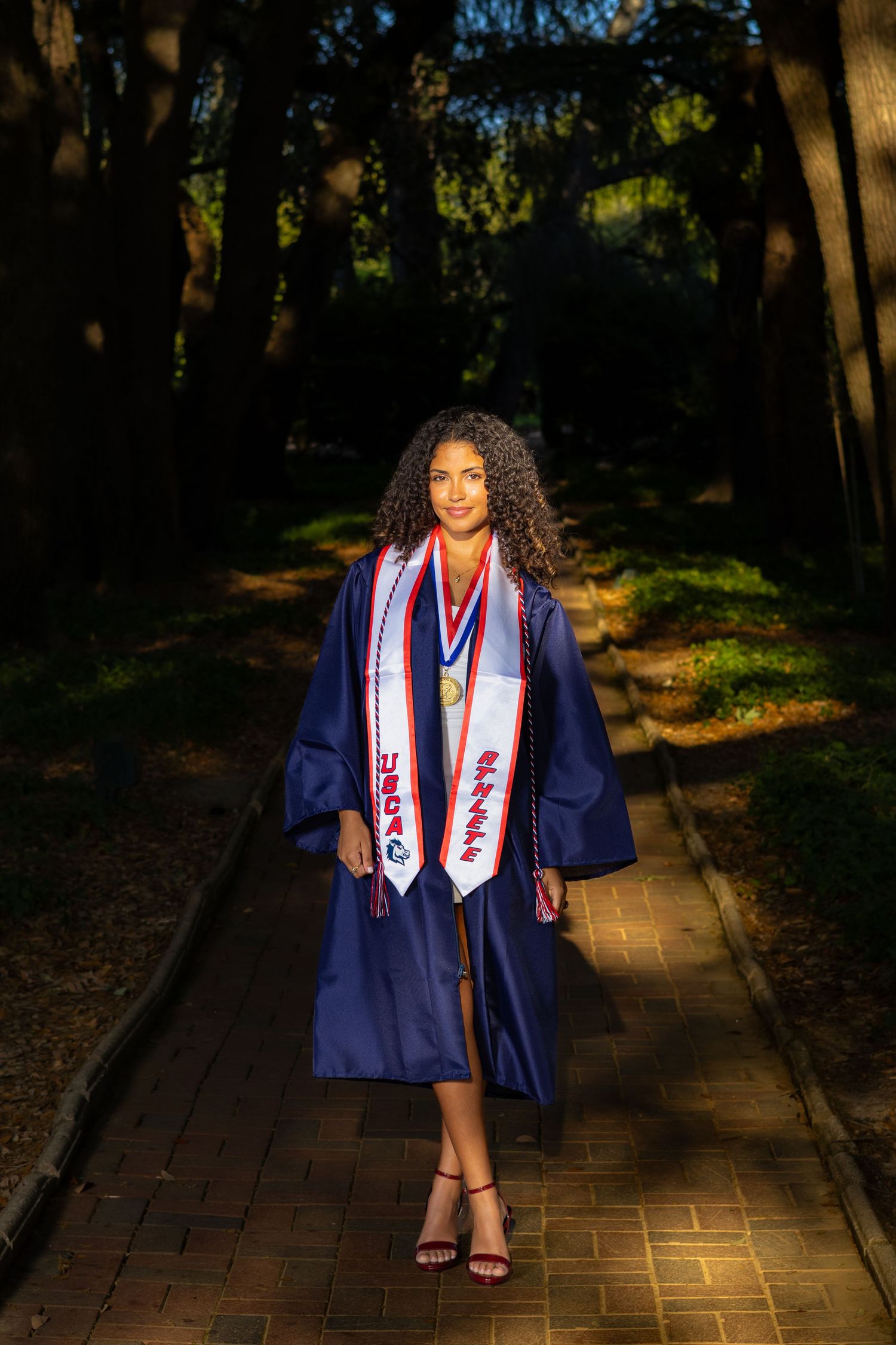 Young woman in blue graduation gown with honor stoles walks a brick path outdoors during golden hour.