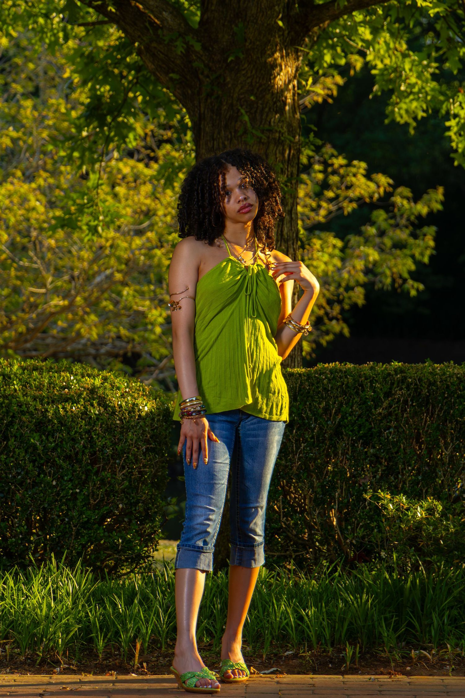 Young Black woman in green halter top and rolled jeans poses outdoors near a large tree with lush green hedges.
