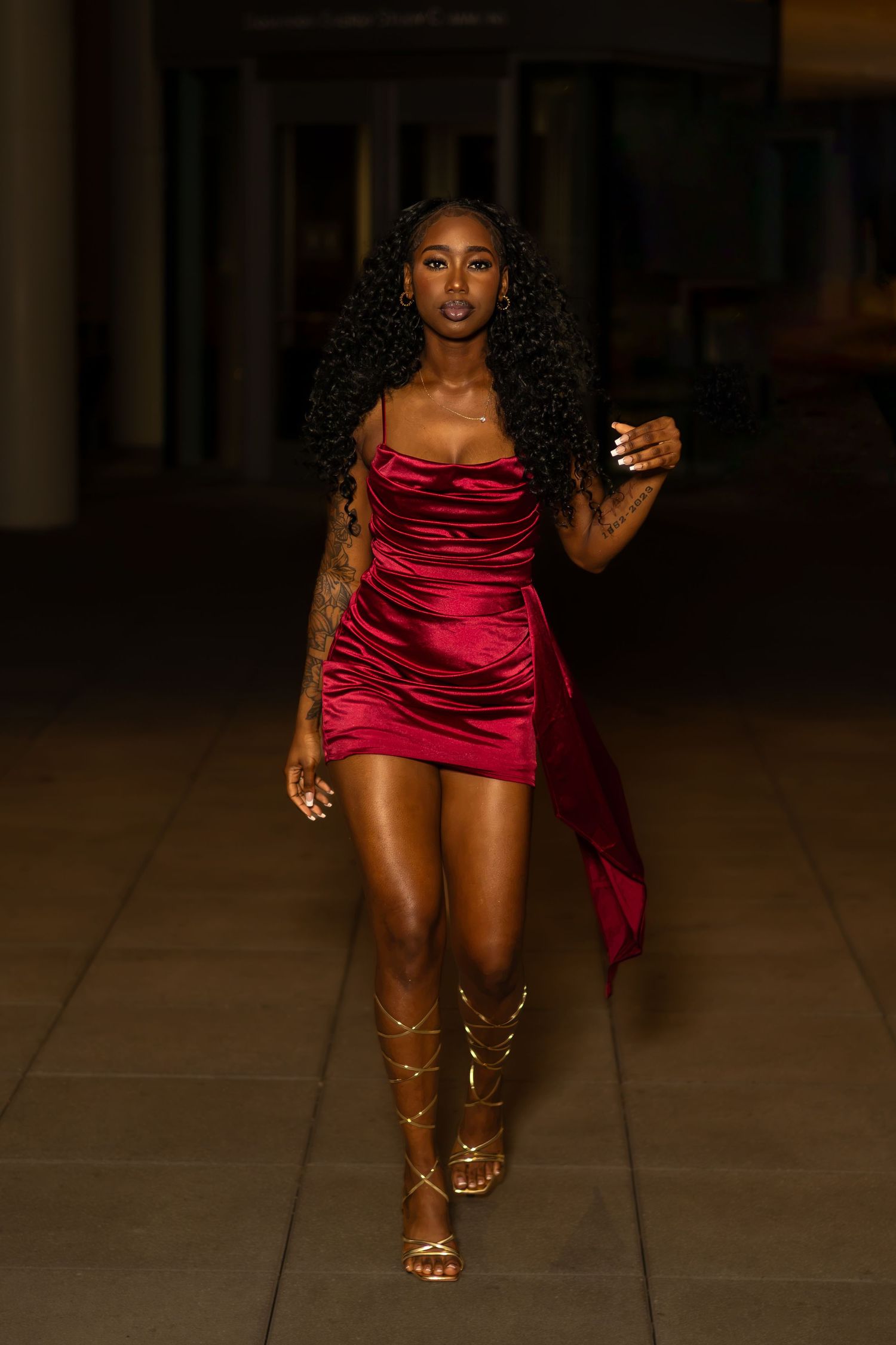 Woman in red satin mini dress and gold gladiator sandals posing confidently at night.