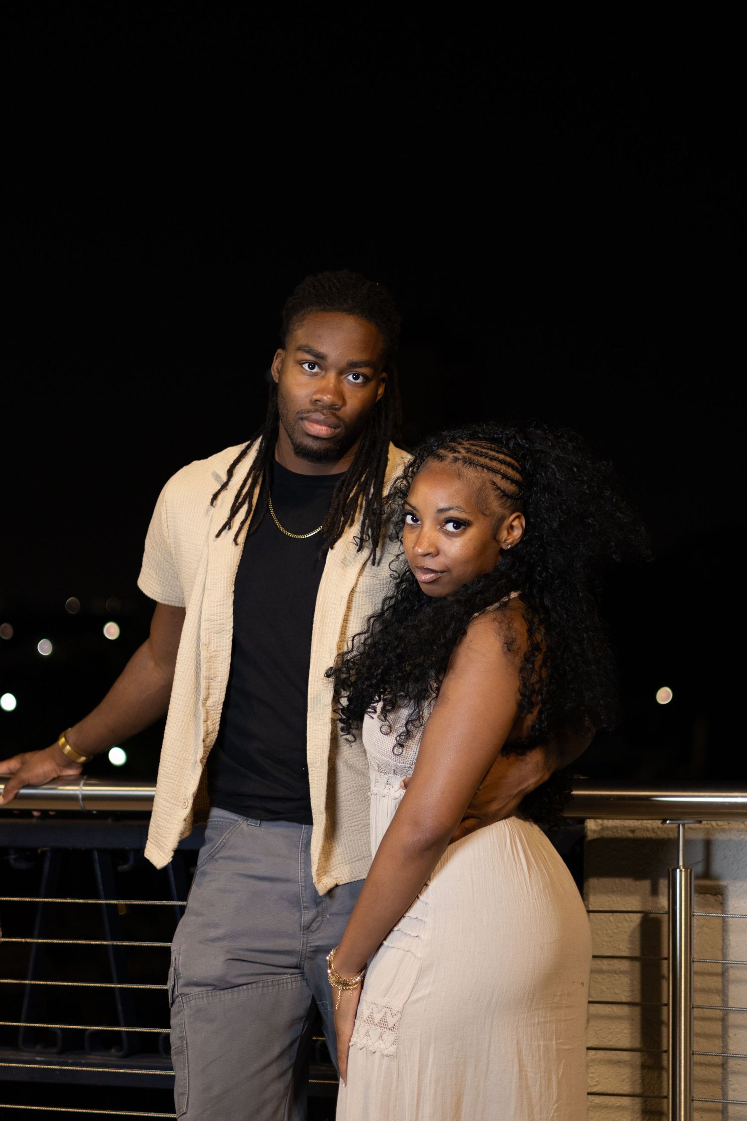 A young Black couple poses elegantly at night on a balcony, dressed in formal attire against a dark skyline.