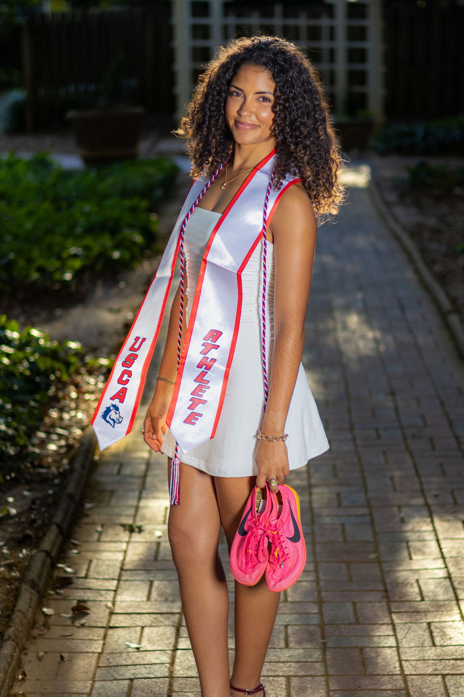 Graduating woman in white dress wearing USCA Athlete sash holds pink sneakers on a brick pathway.
