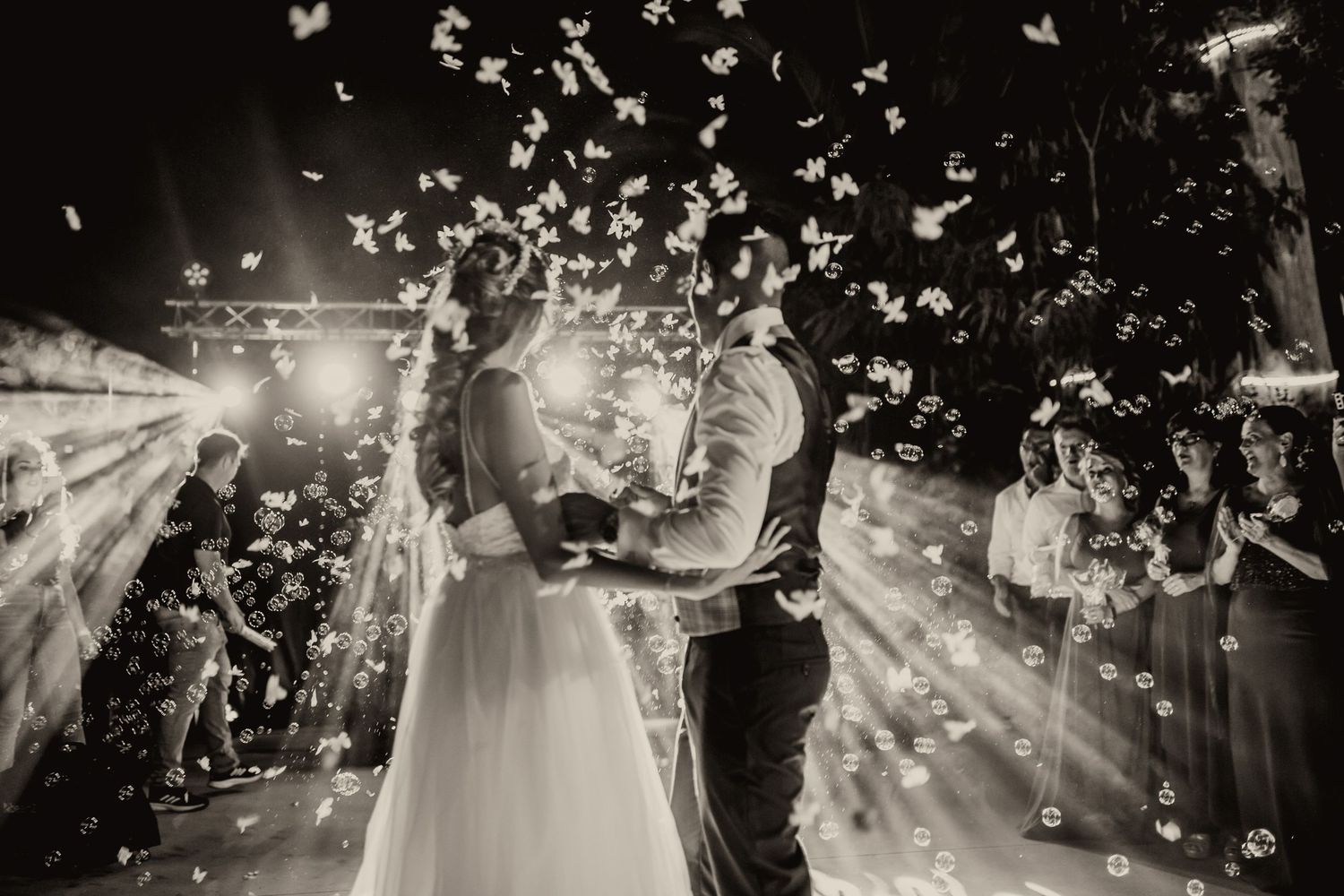 Foto de boda en blanco y negro de pareja compartiendo el primer baile rodeado de pétalos de flores que caen y luces brillantes.