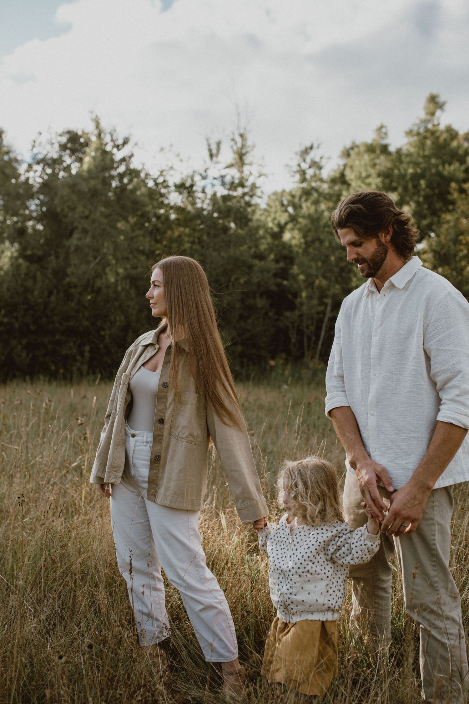 A young family of three walks through a golden meadow, parents holding their toddler's hands near lush green trees. Family Photography.