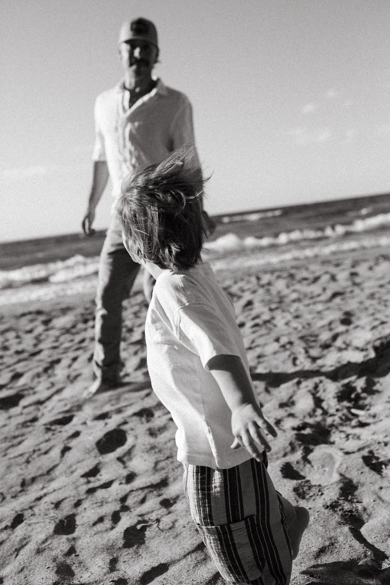 Black and white photo of a child running toward a man on a sandy beach with ocean waves in the background with Dad. Family Photography.