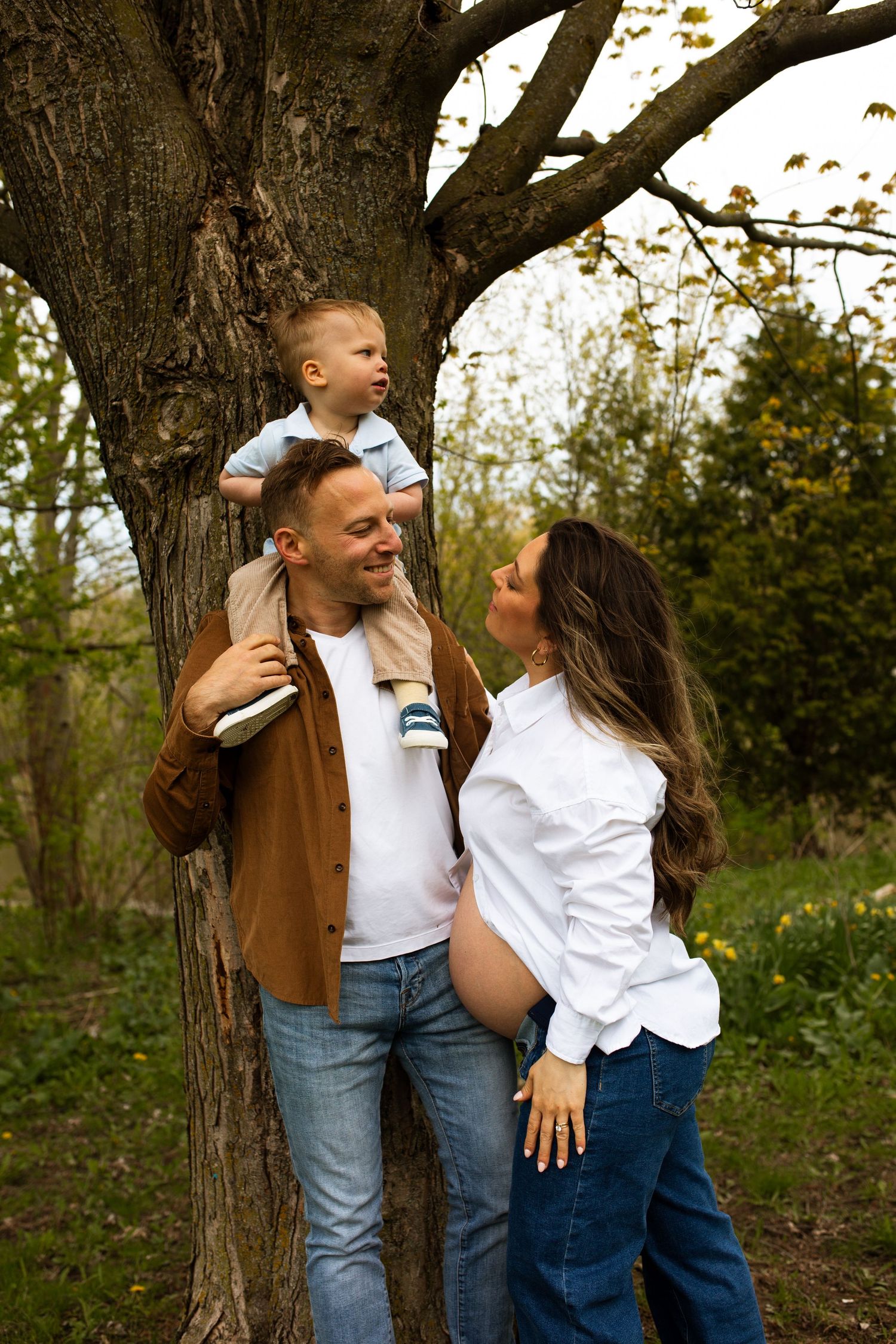 Happy family outdoors, pregnant mother in white shirt, father carrying toddler on shoulders by a tree. Maternity Photography.