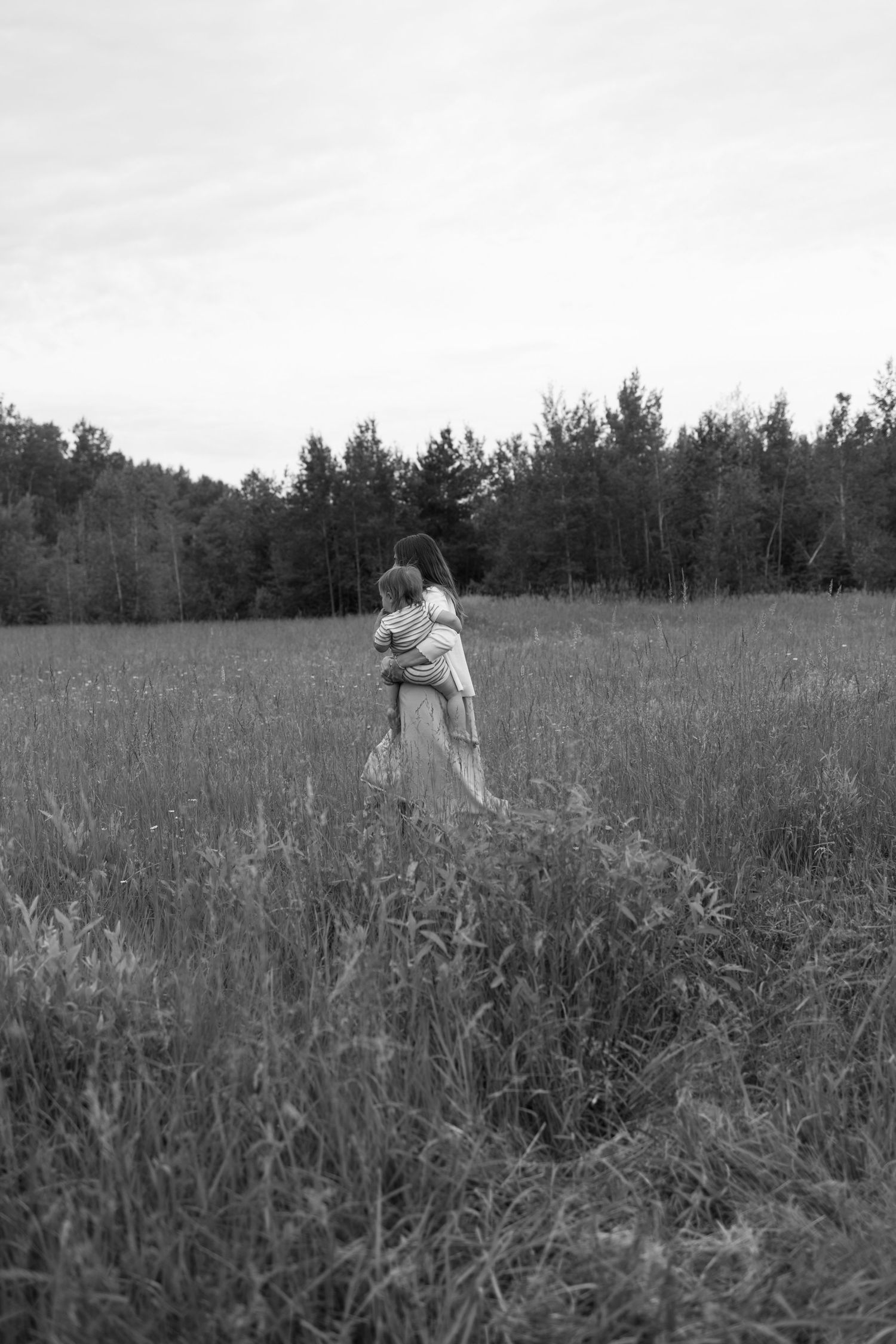 Black and white photo of a woman holding a child in a lush meadow with a forest backdrop. Family Photography.