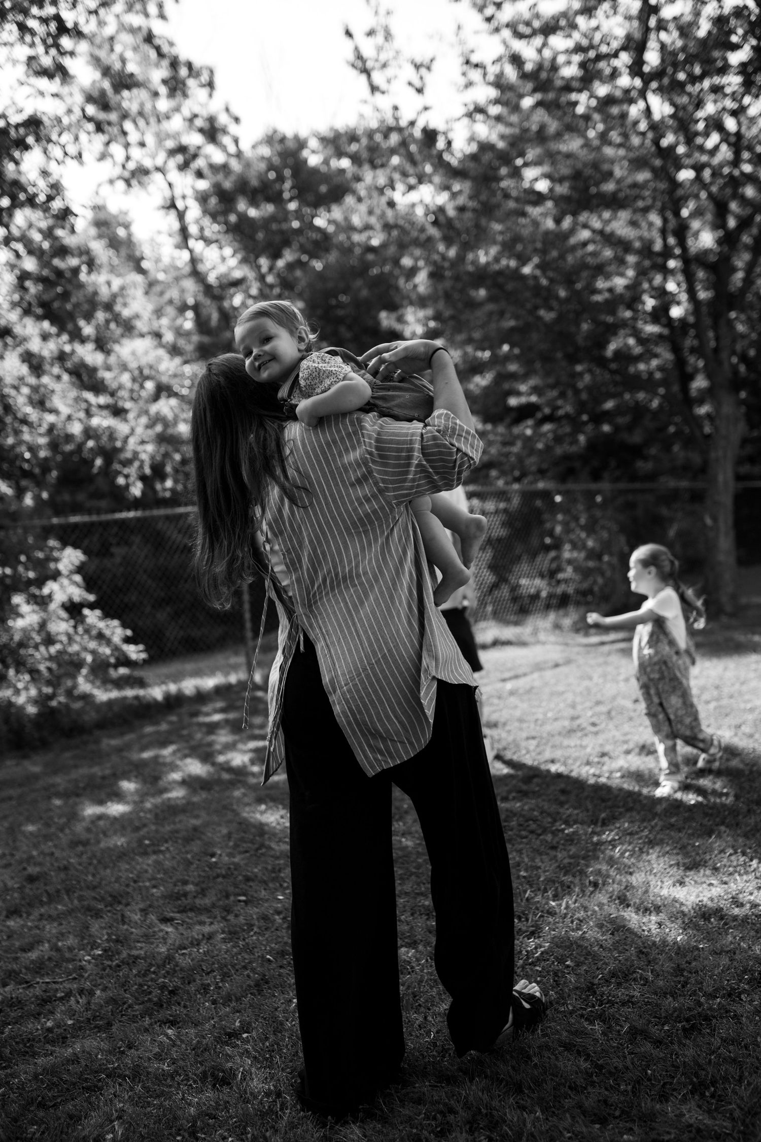 Black and white photo of a mother lifting her child in a backyard while another child plays in the background. Family Photography.