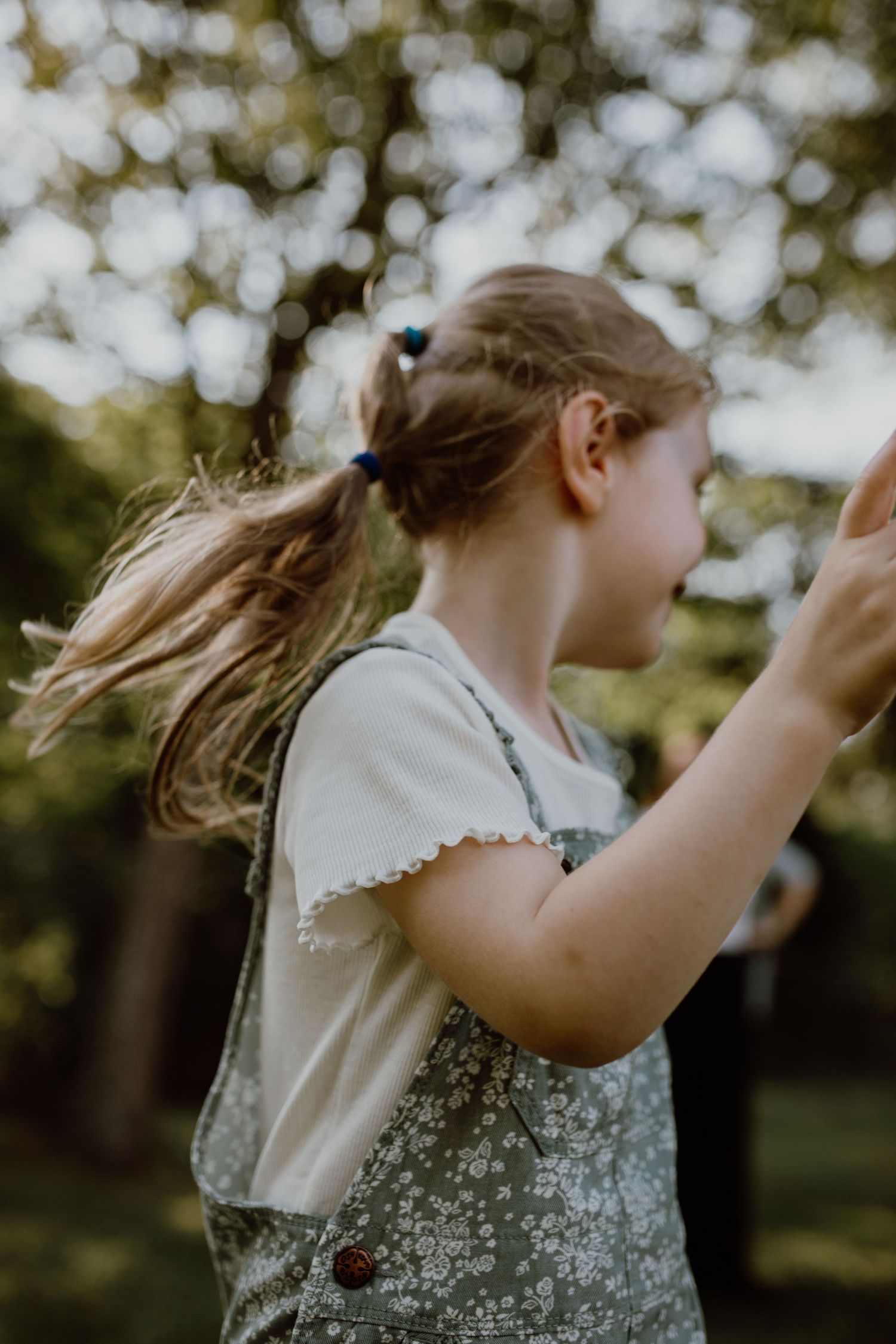 A young girl with a ponytail wearing floral overalls plays outdoors in a sunlit garden. Family Photography.