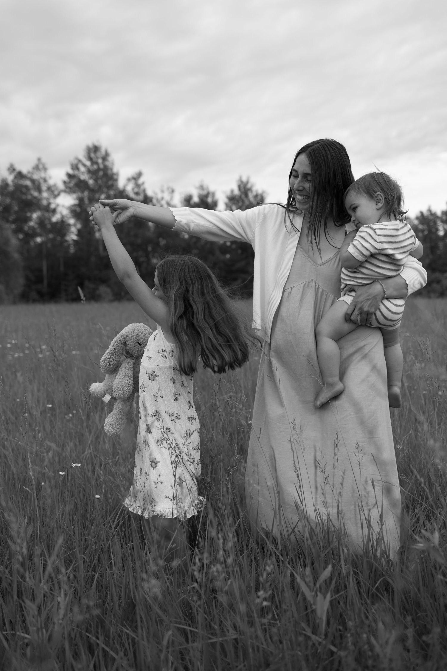 Black and white photo of a mother holding a baby while dancing with her young daughter in a field. Family Photography.
