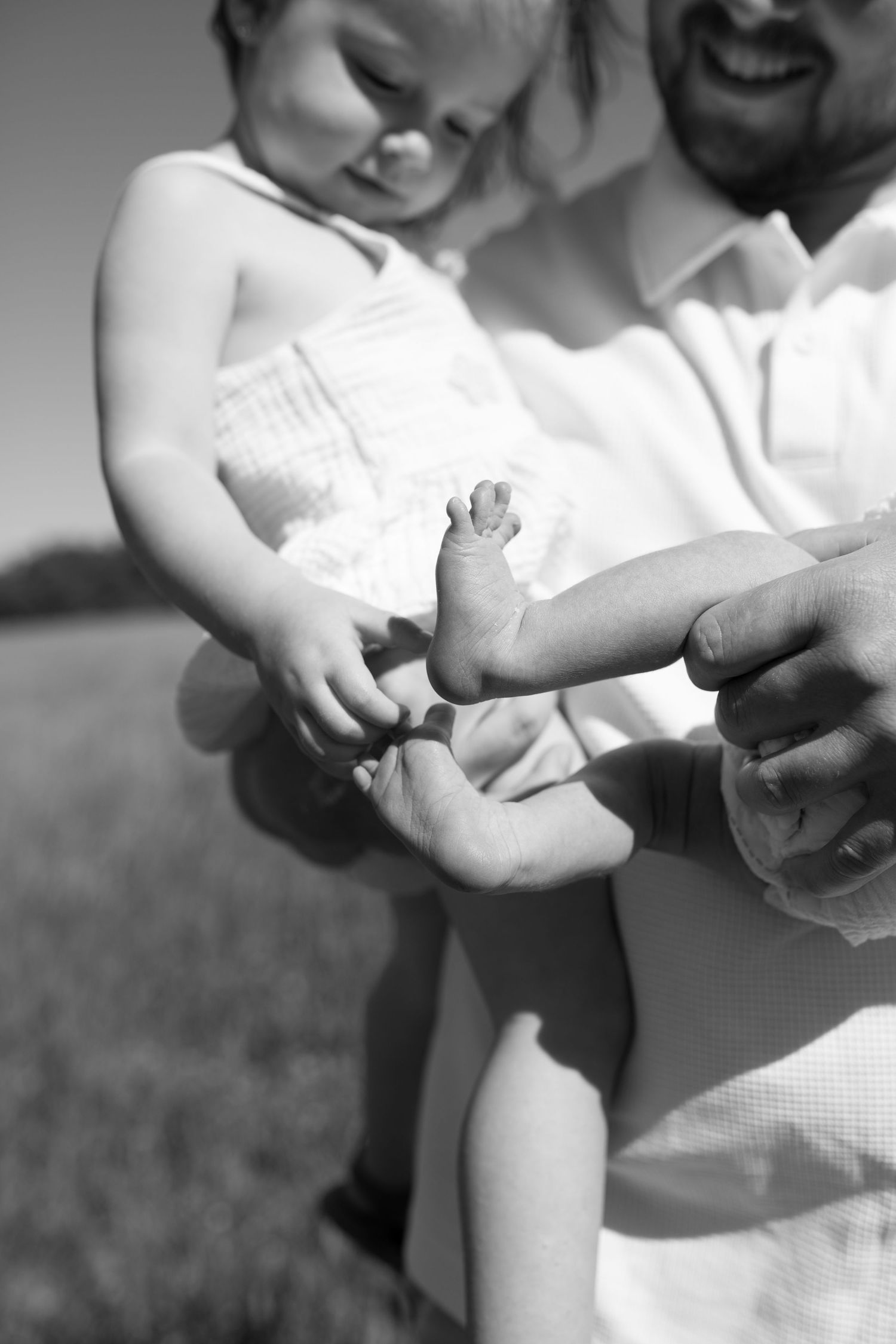 Black and white photo of a father holding his baby, gently touching the infant's tiny foot outdoors. Family Photography. Newborn Photography