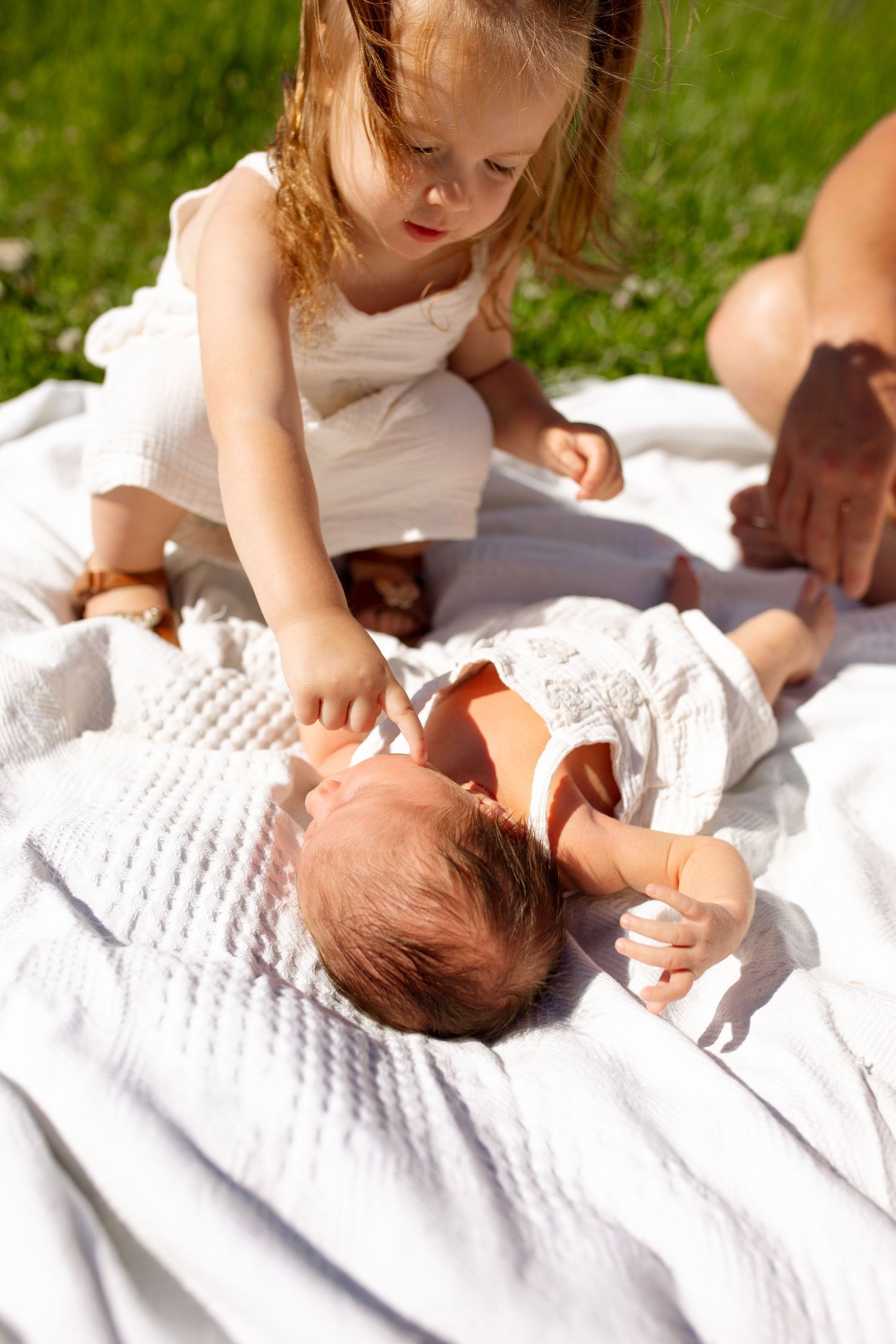 Young girl in white dress gently touches newborn baby lying on white blanket outdoors on sunny day. Family Photography. Newborn Photography.