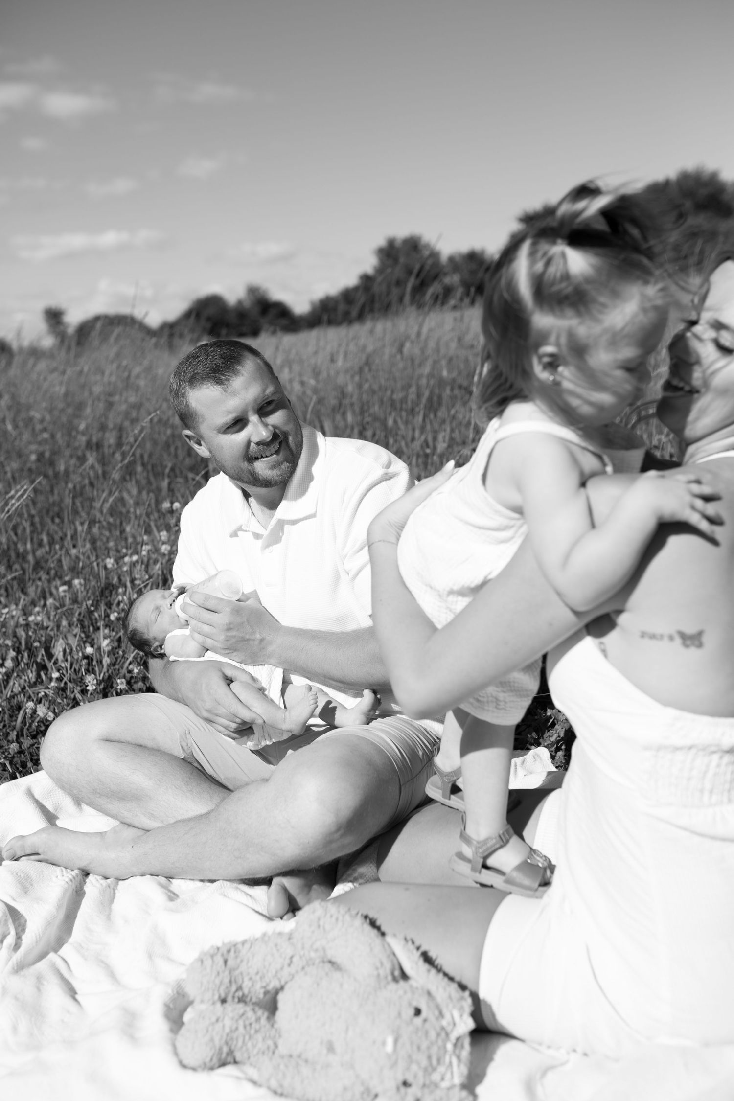 Black and white photo of a family enjoying a picnic in a field, father holding a newborn baby with children nearby. Family Photography.