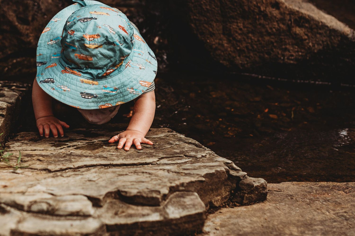 A toddler in a colorful bucket hat exploring rocky terrain near a natural water feature.