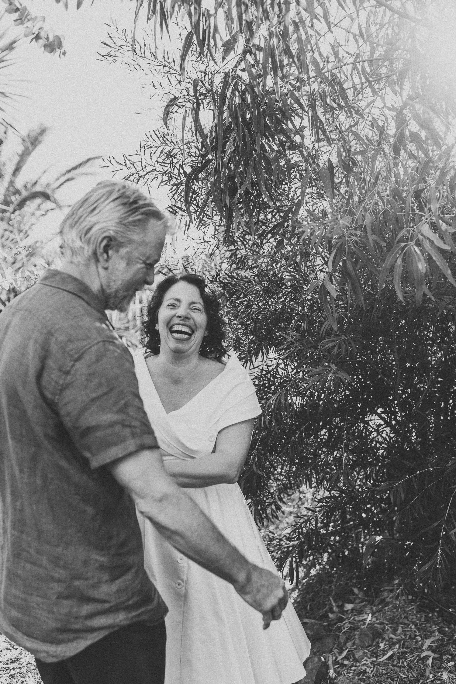 Black and white photo of a bride laughing with groom while holding hands in a garden.