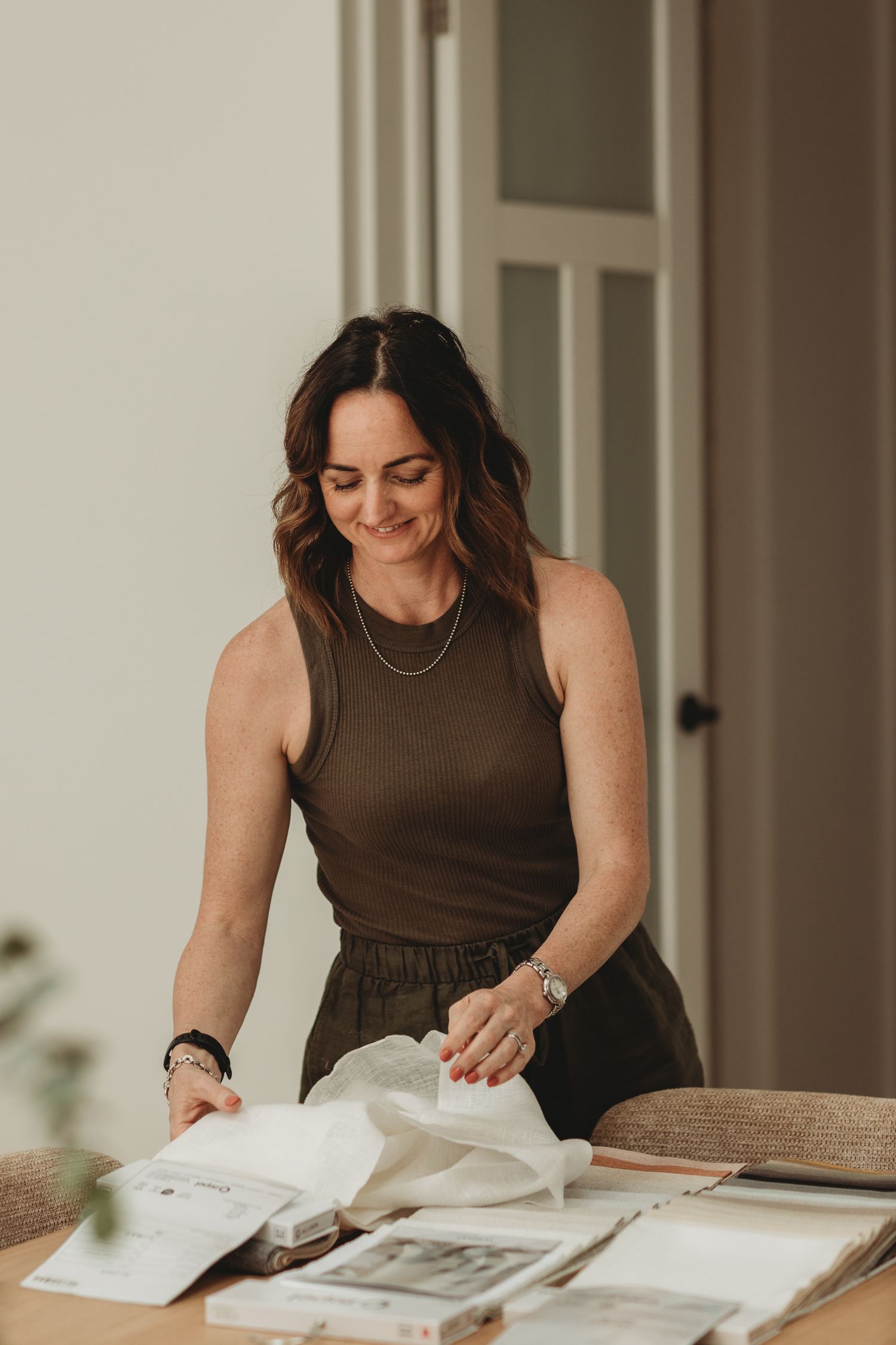 Smiling brunette woman in olive outfit browsing fabric samples and design books at a wooden table.