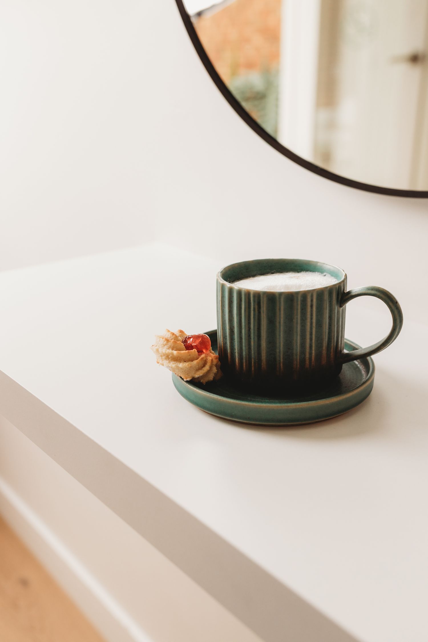 Green ribbed ceramic mug of frothy latte on a matching saucer with a jam-topped butter cookie, on a white shelf beside a round mirror.