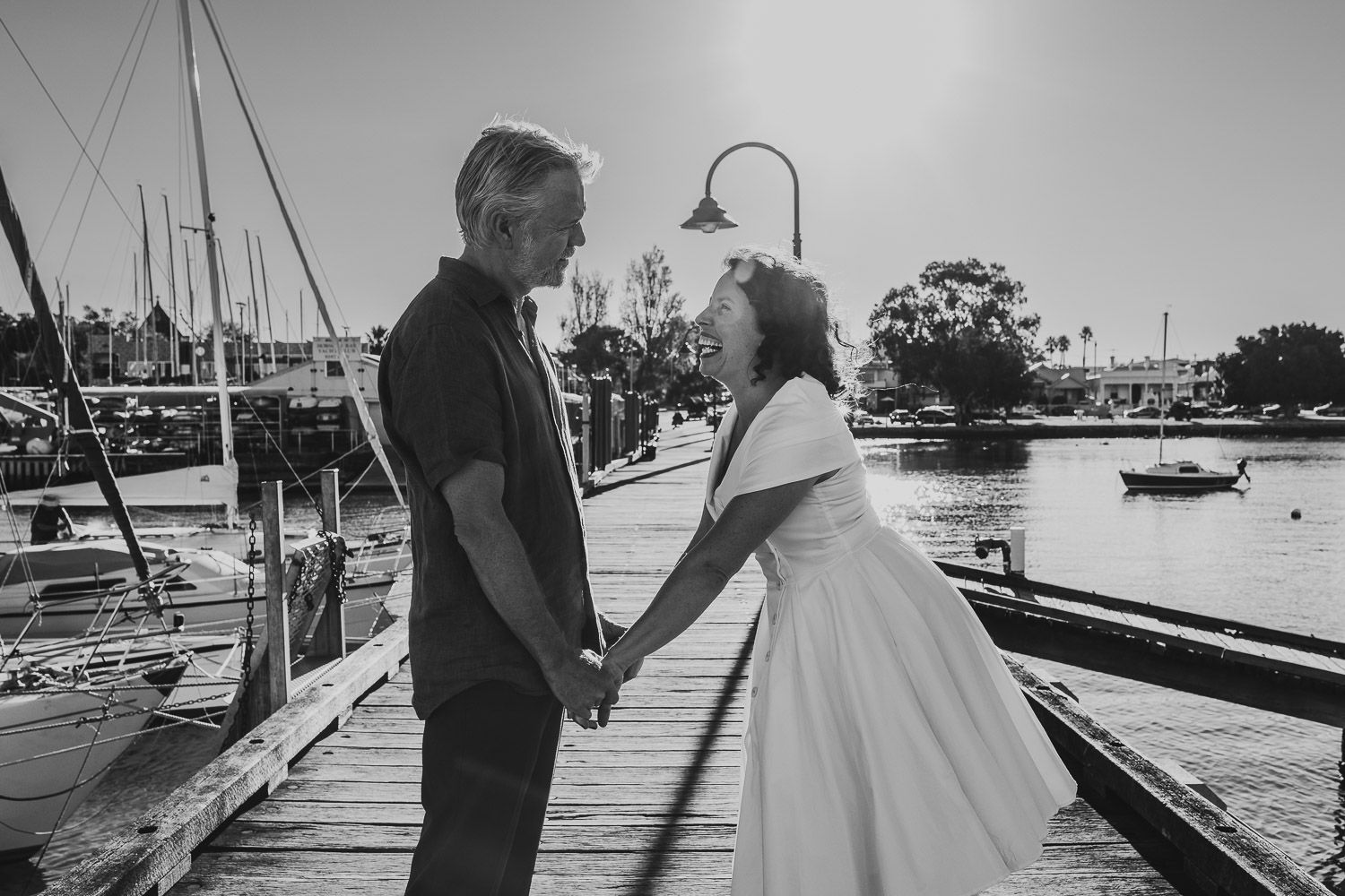 Bride and groom holding hands and smiling at each other on a wooden pier at a marina in black and white.