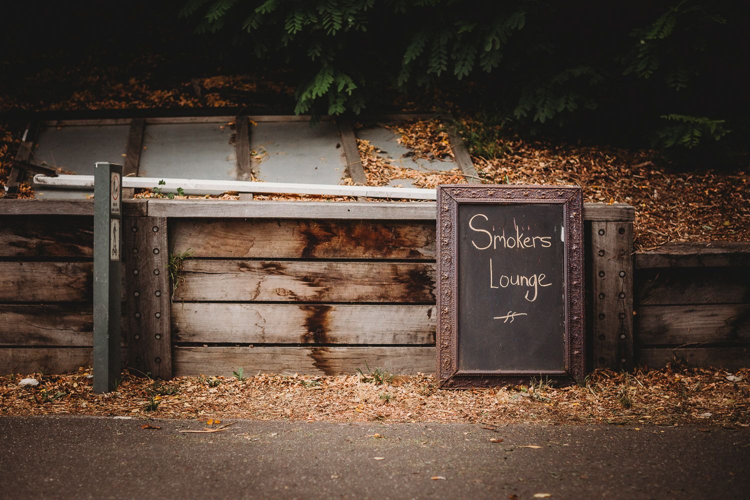 Rustic chalkboard sign reading Smokers Lounge leaning against a weathered wooden fence surrounded by autumn leaves.