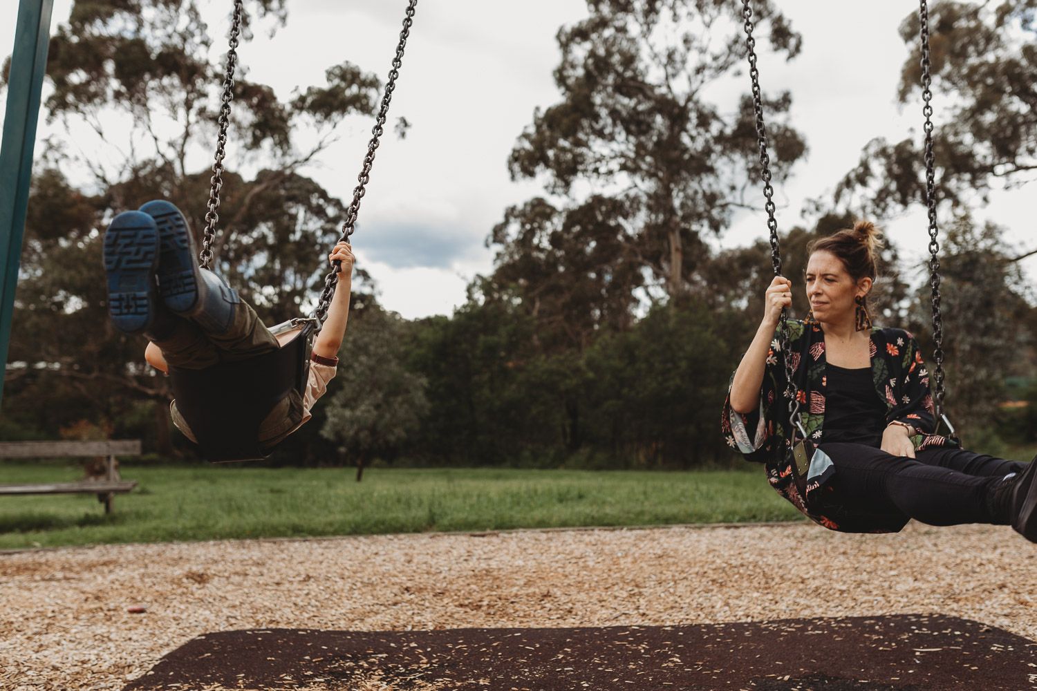 Mother and child enjoying swings together at a park surrounded by tall trees on cloudy day.