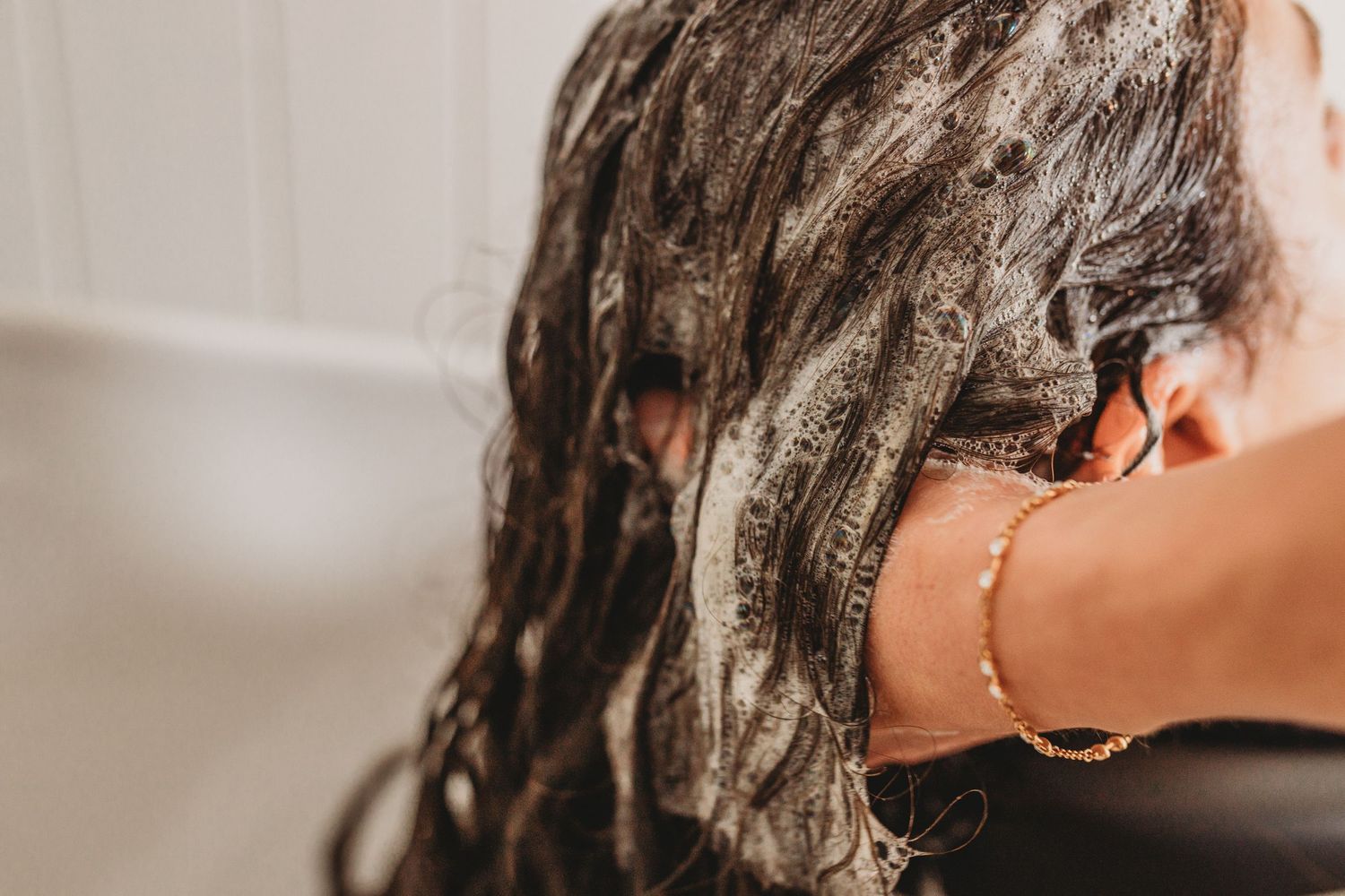 Woman with long dark wet hair covered in shampoo lather washing her hair in a bright bathroom, wearing a gold bracelet.