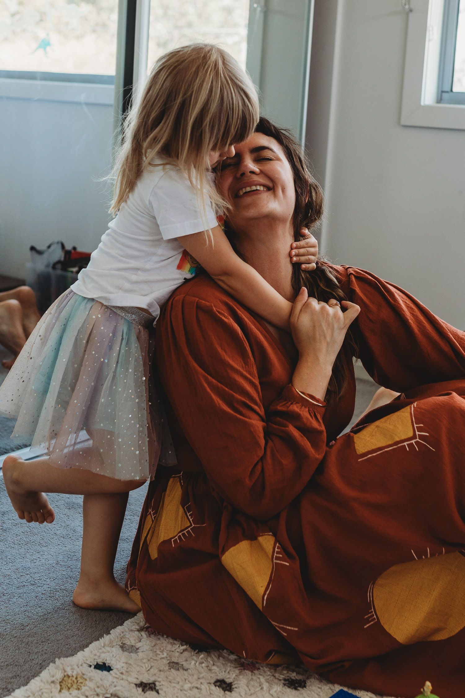 Laughing mother in rust robe being hugged by young daughter in tutu skirt on carpet floor.