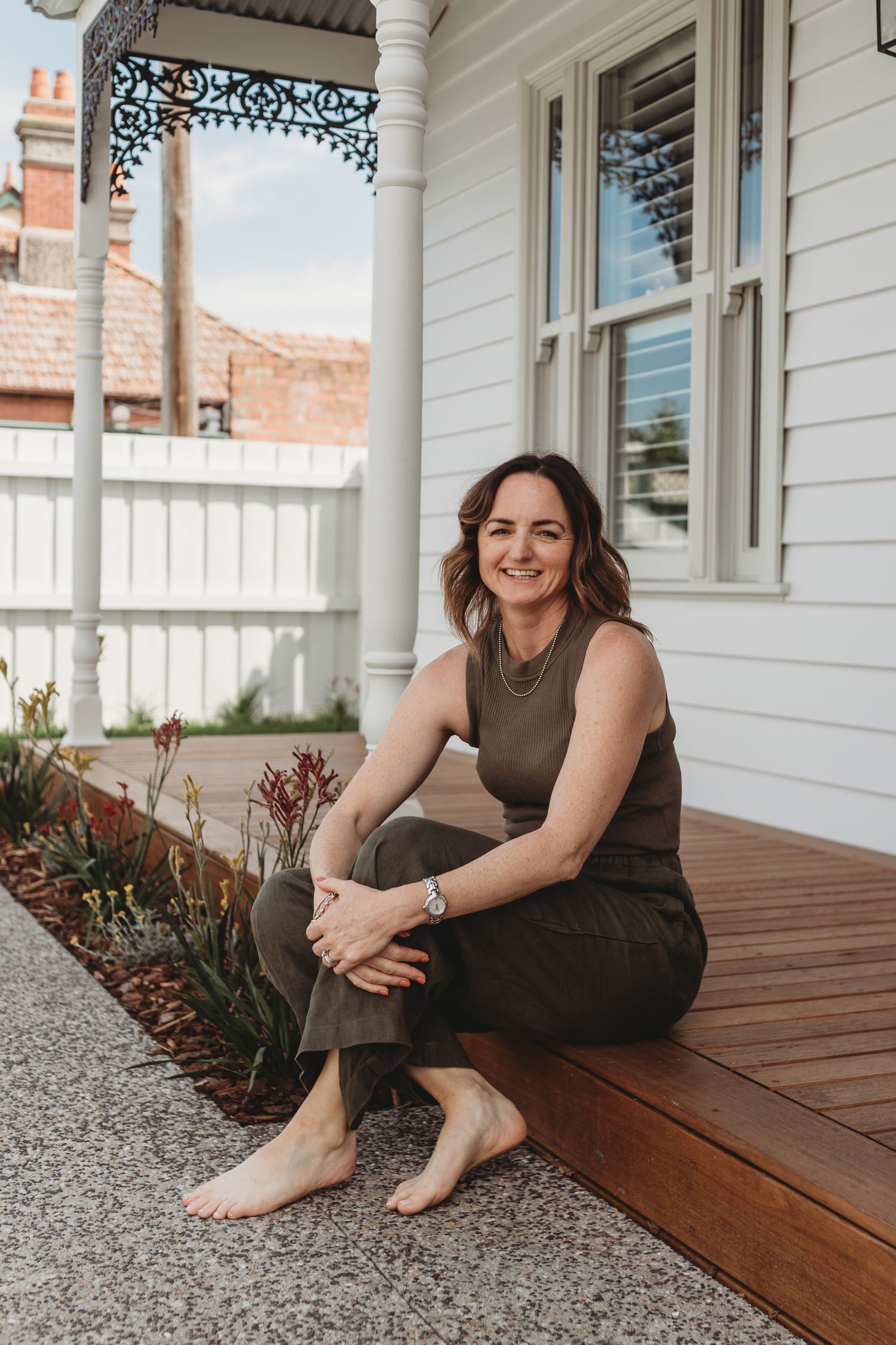 Smiling brunette woman sitting barefoot on a timber deck of a classic white weatherboard Victorian home.