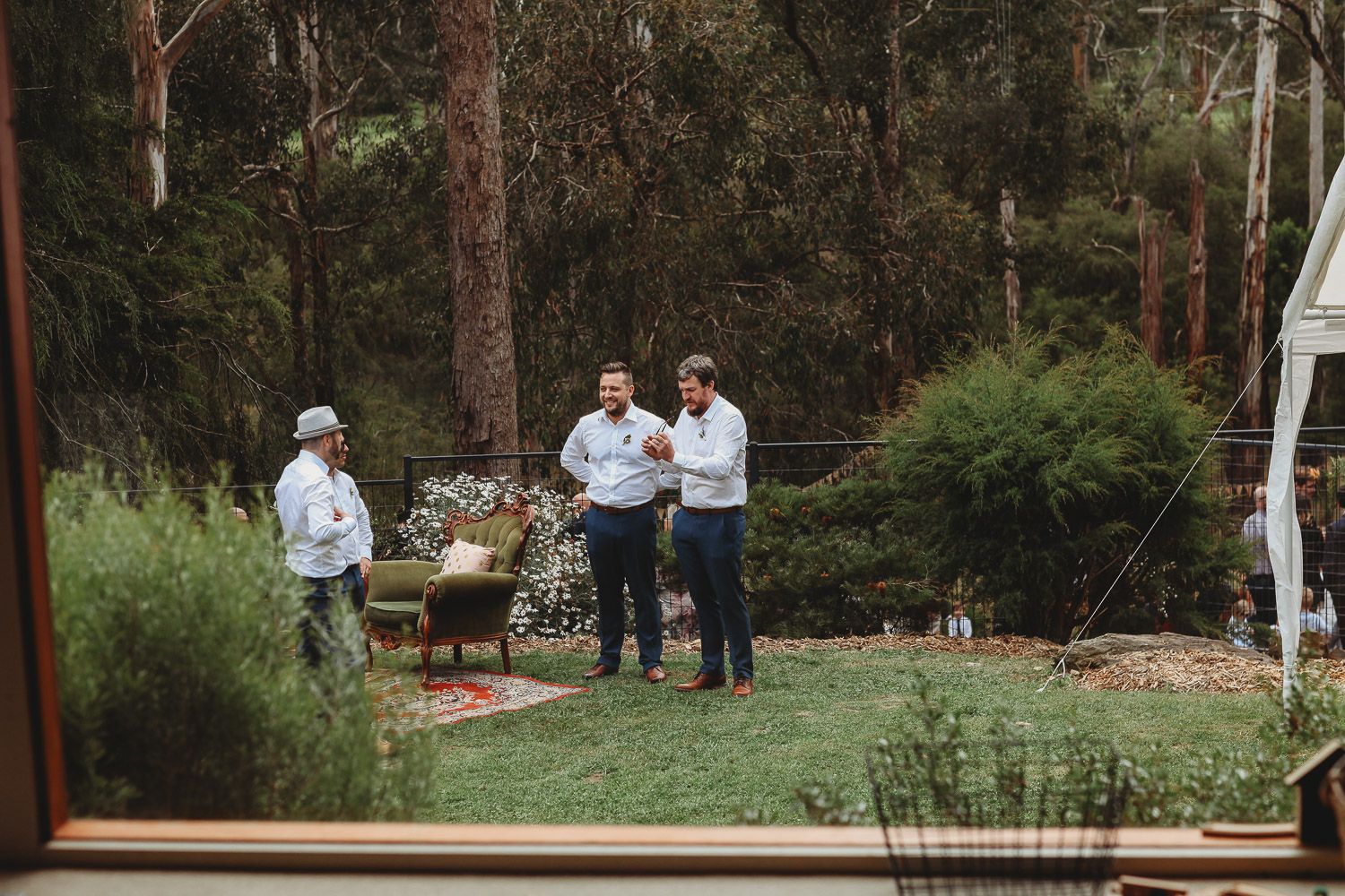 Three men conversing in lush garden setting viewed through glass window.