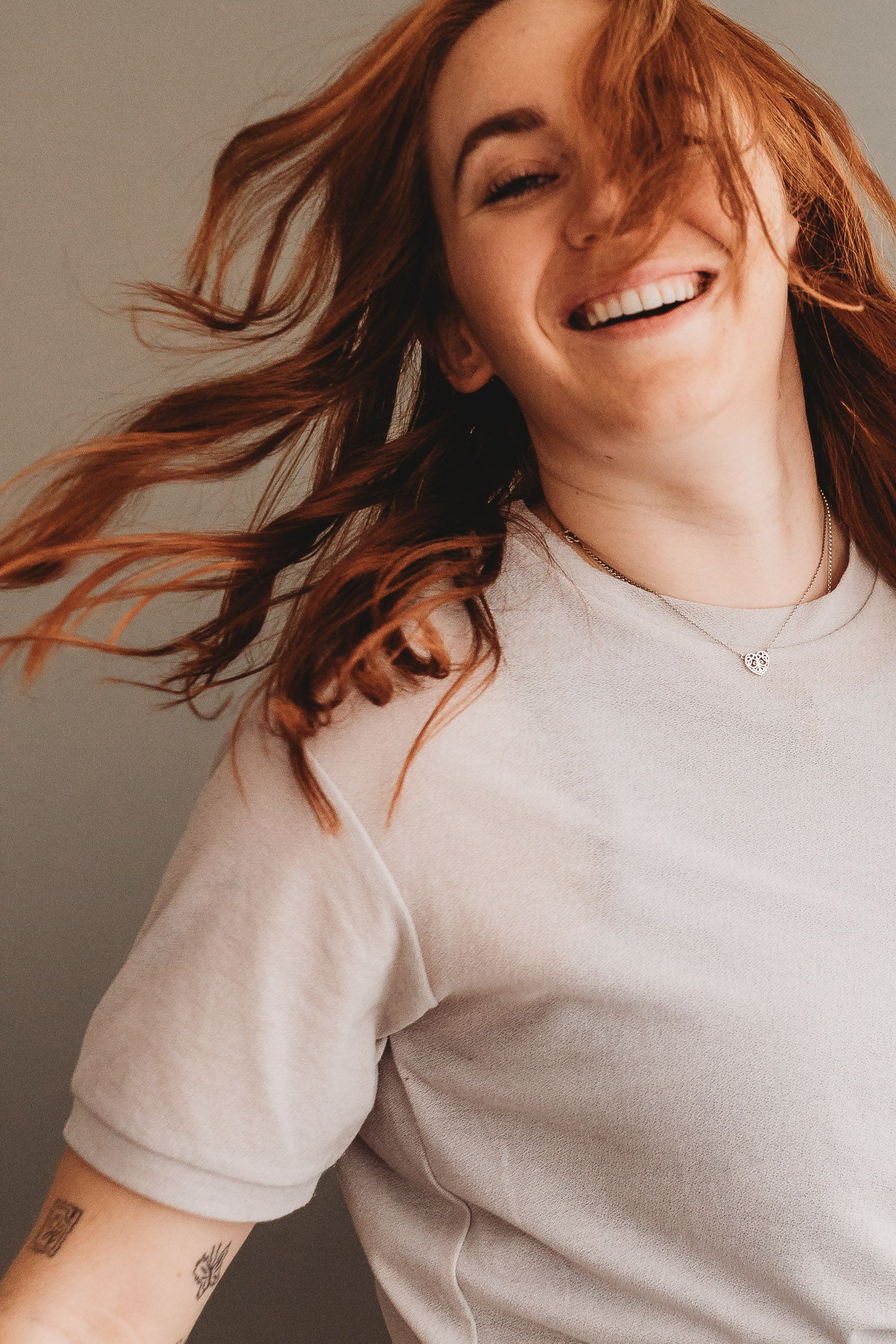 Happy redhead woman with flowing hair laughing joyfully wearing a light gray t-shirt with arm tattoo.