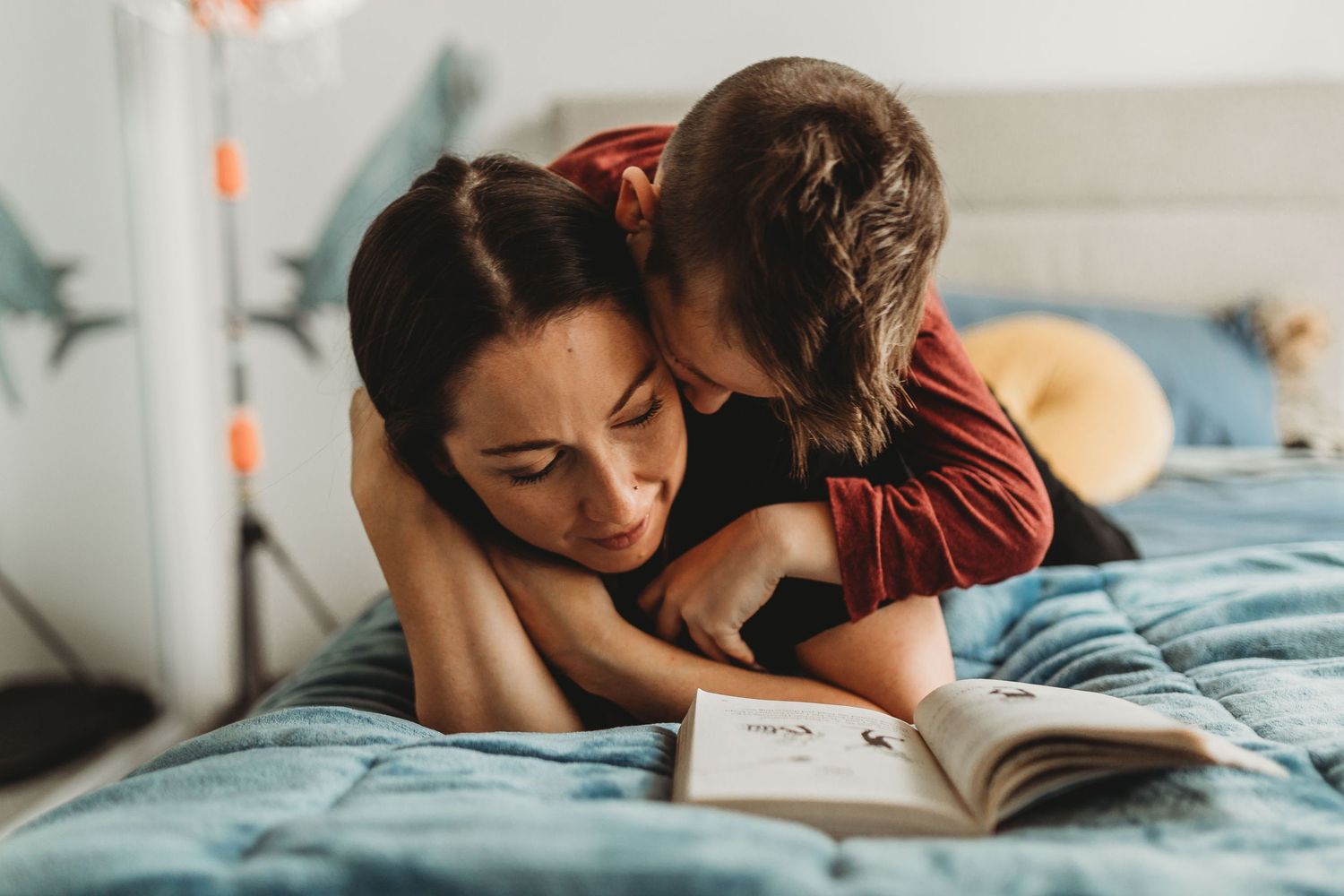 A child lovingly embraces his mother while she reads a book on a blue bed.