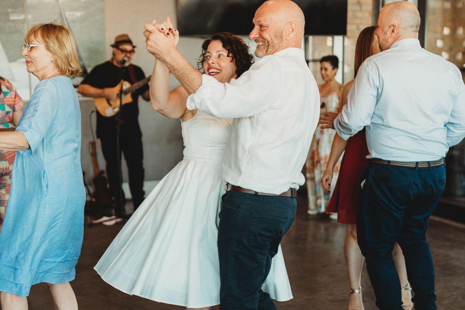 Guests dancing joyfully at a lively wedding reception with a live guitarist performing in the background.