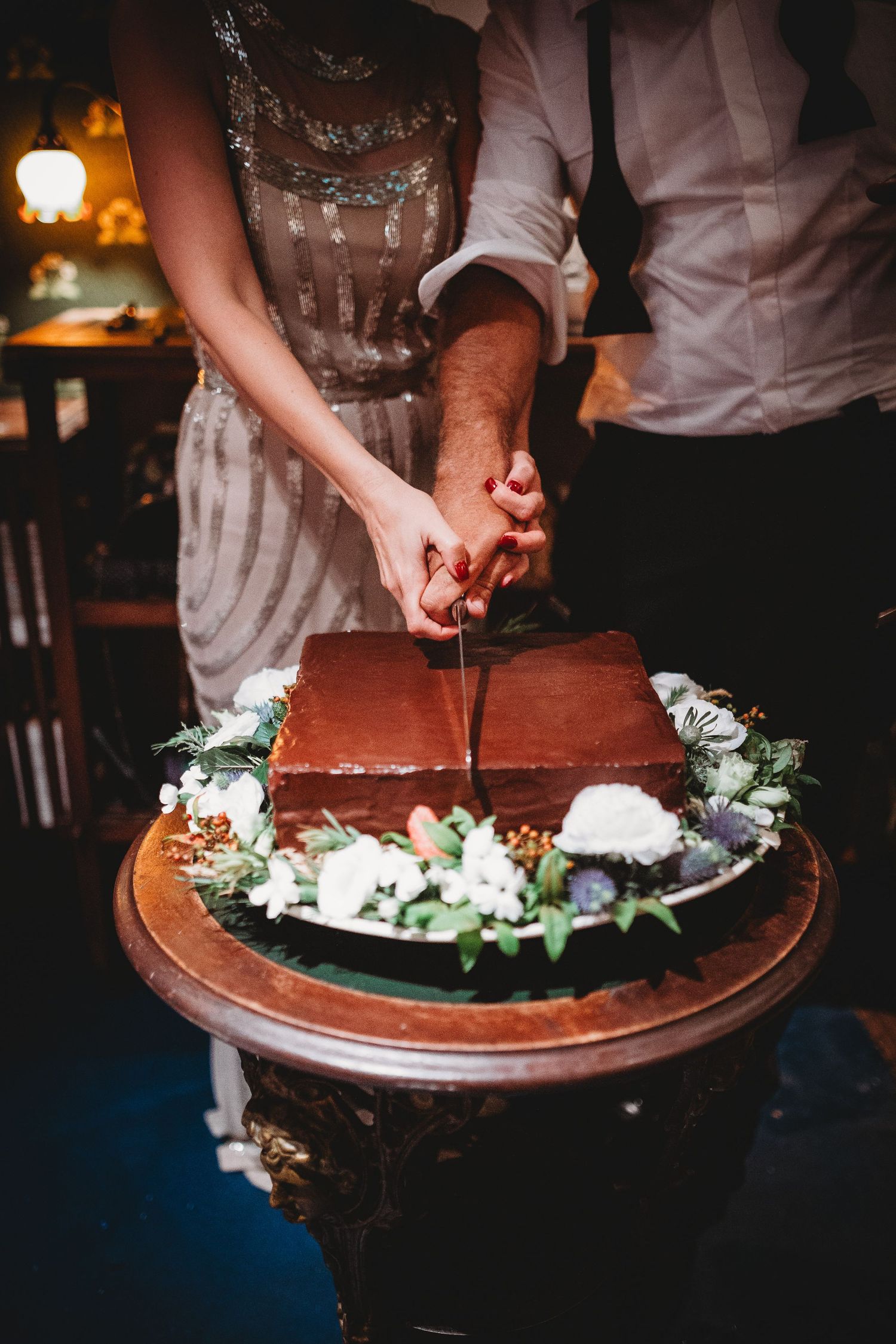 A bride and groom cutting a square chocolate wedding cake adorned with white flowers.