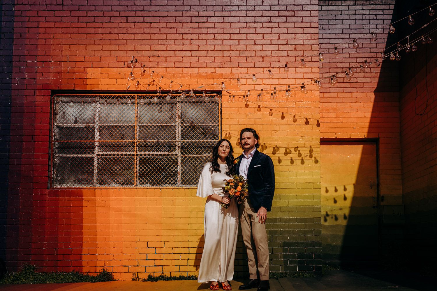 A bride and groom standing together in front of a vibrant rainbow-colored brick wall.