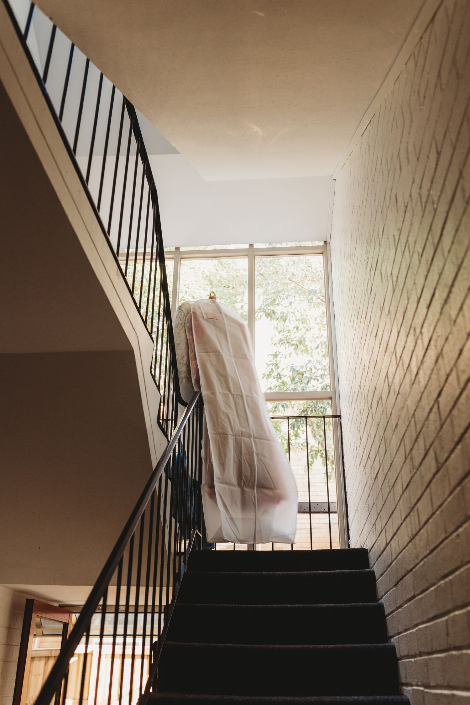Wedding dress in garment bag hanging on staircase railing near window.