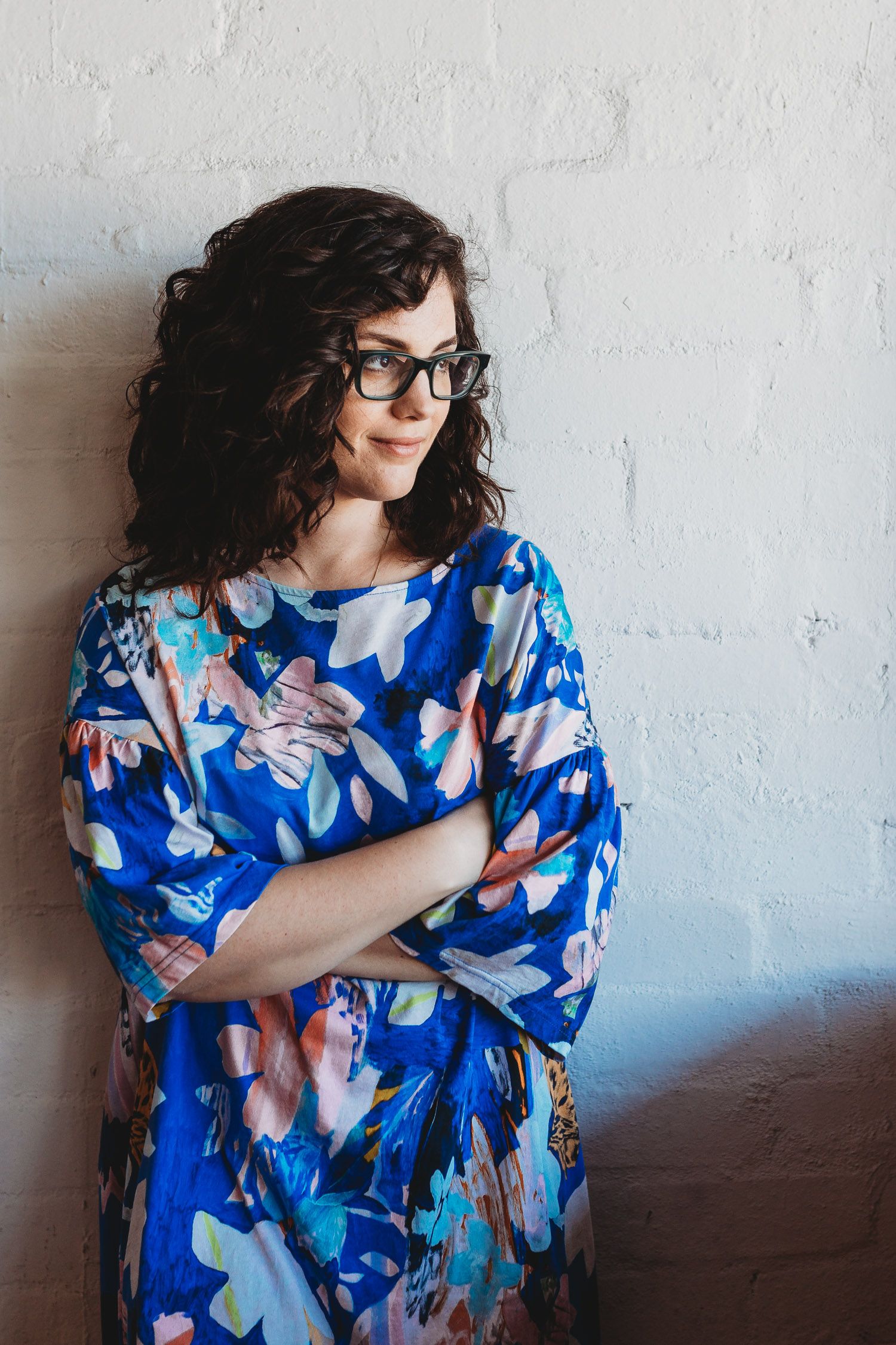 A woman with curly hair and glasses in a blue floral dress leans against a white brick wall.