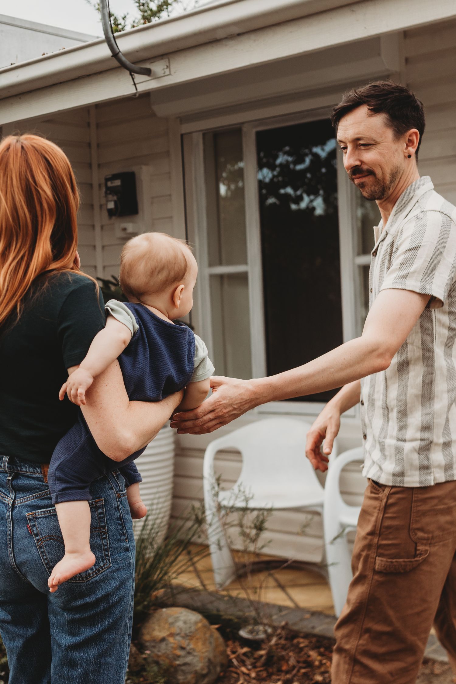 A smiling man in a striped shirt reaches out to a baby held by a red-haired woman on the front porch of a home.