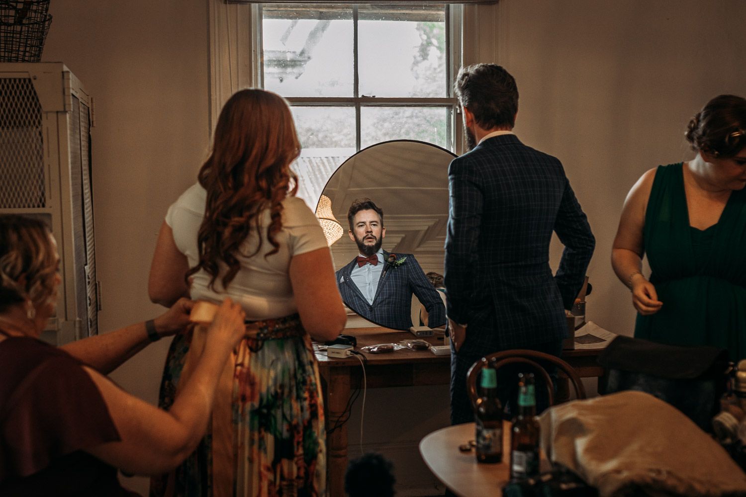 A groom getting ready with friends gathered around a mirror in a bright room.