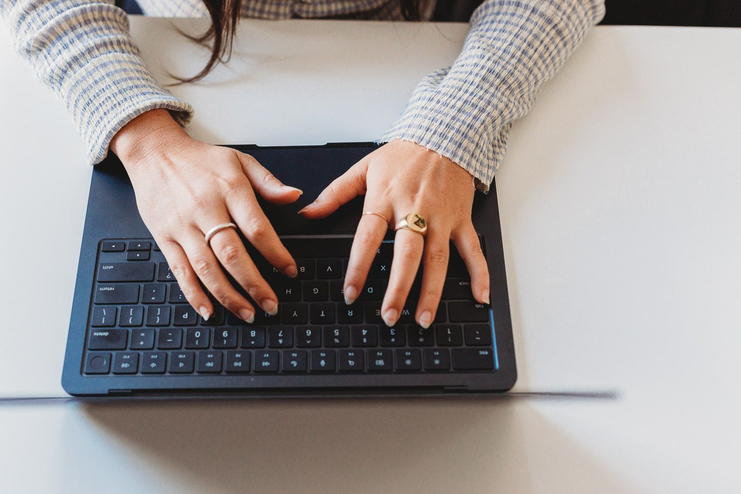 Woman wearing gold rings typing on a black wireless keyboard on a white desk.