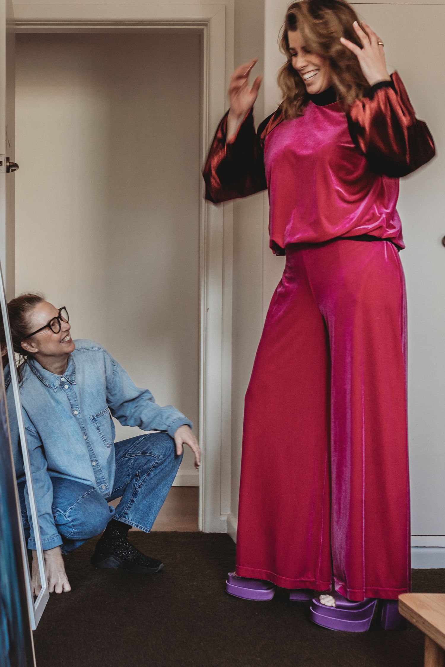 Woman in pink satin wide-leg pantsuit laughing while designer adjusts hem on floor.