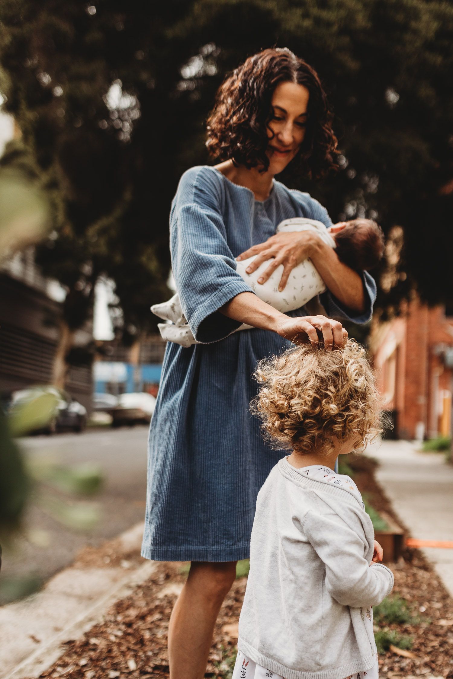 A curly-haired mother holding a newborn while her toddler stands nearby on an outdoor path.