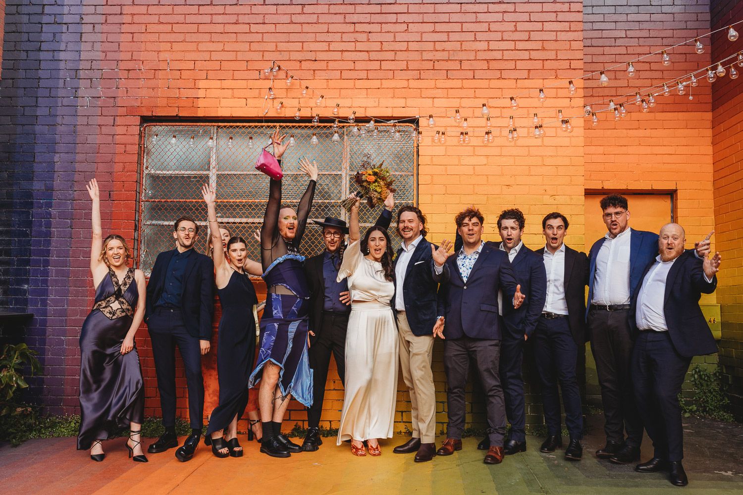 A joyful wedding group photo in front of a colorful mural on a brick wall.
