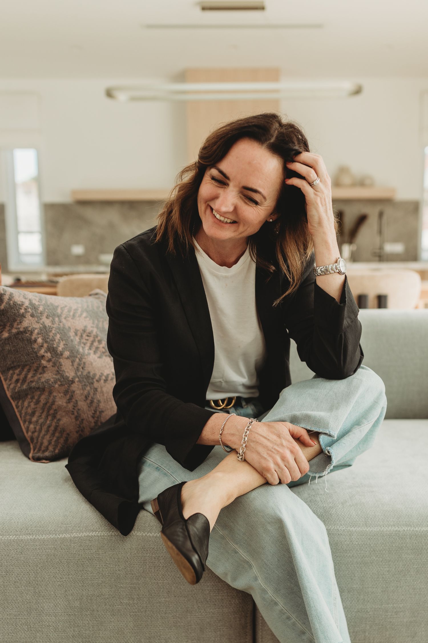 Smiling woman in black blazer and light jeans relaxing on a grey sofa in a modern open-plan living space.
