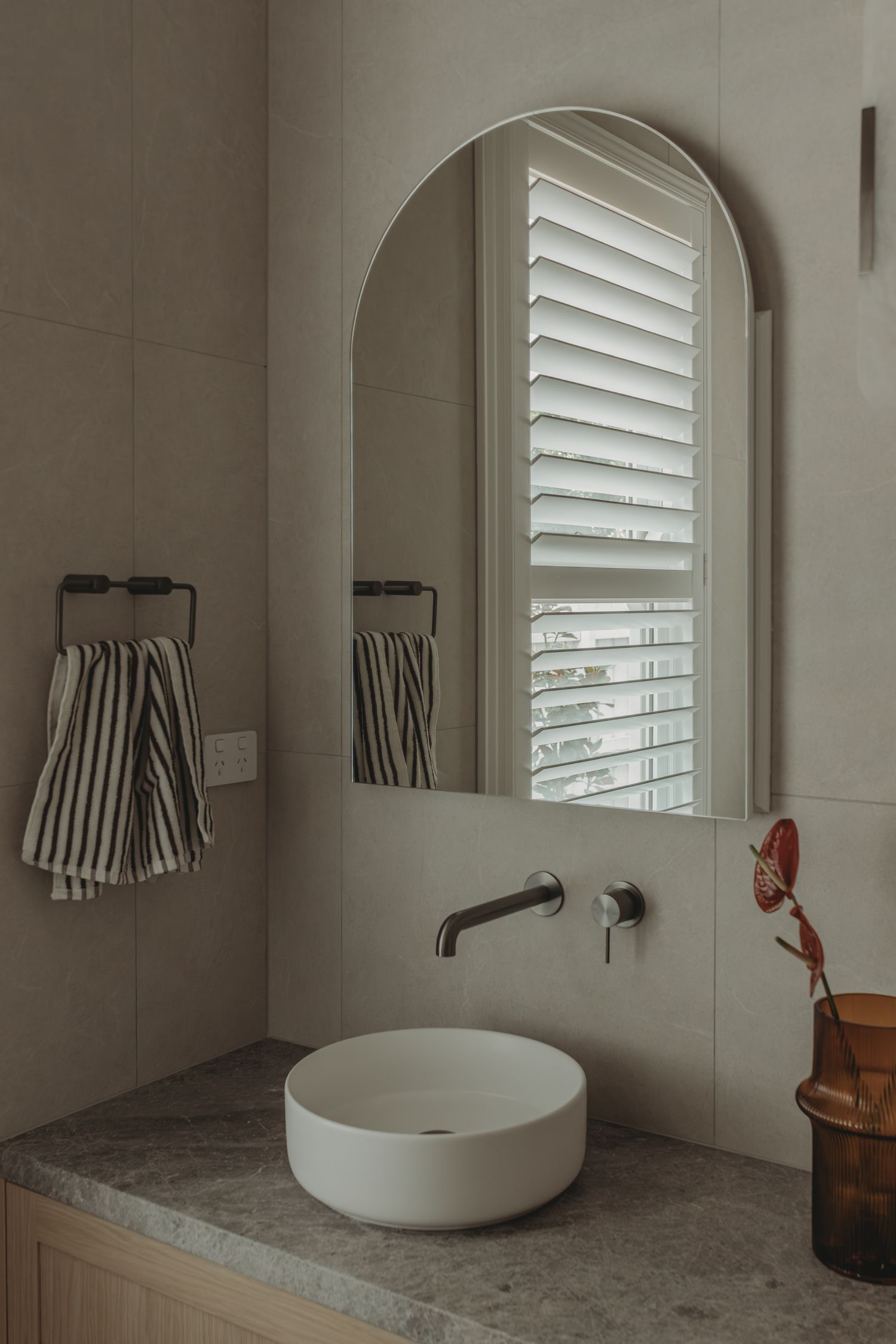 Modern bathroom vanity with round vessel sink, arched mirror, wall-mounted faucet, and striped towels on grey marble countertop.