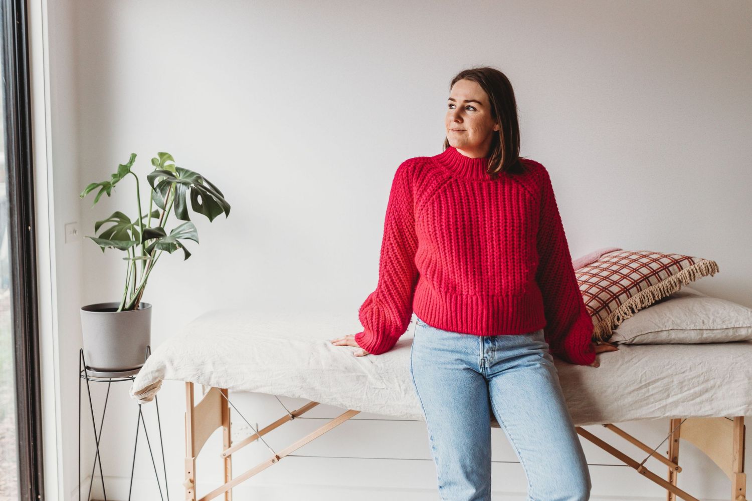 A woman in a red knit sweater and jeans sits on a massage table beside a potted plant.