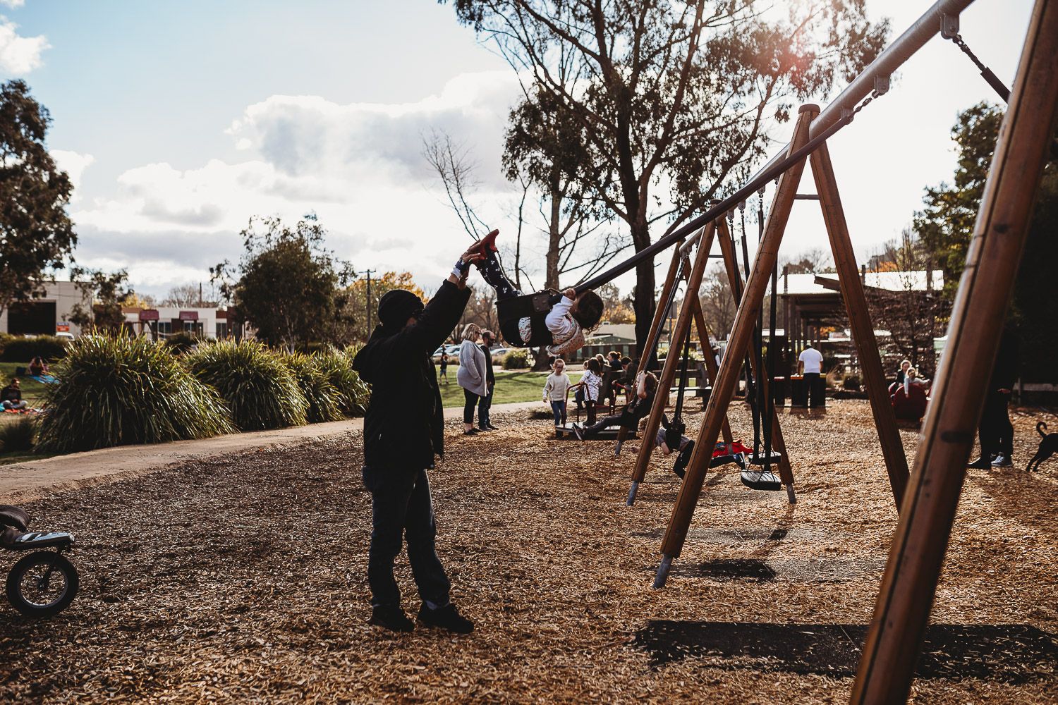 Children play on wooden swings at a sunny outdoor playground surrounded by trees.